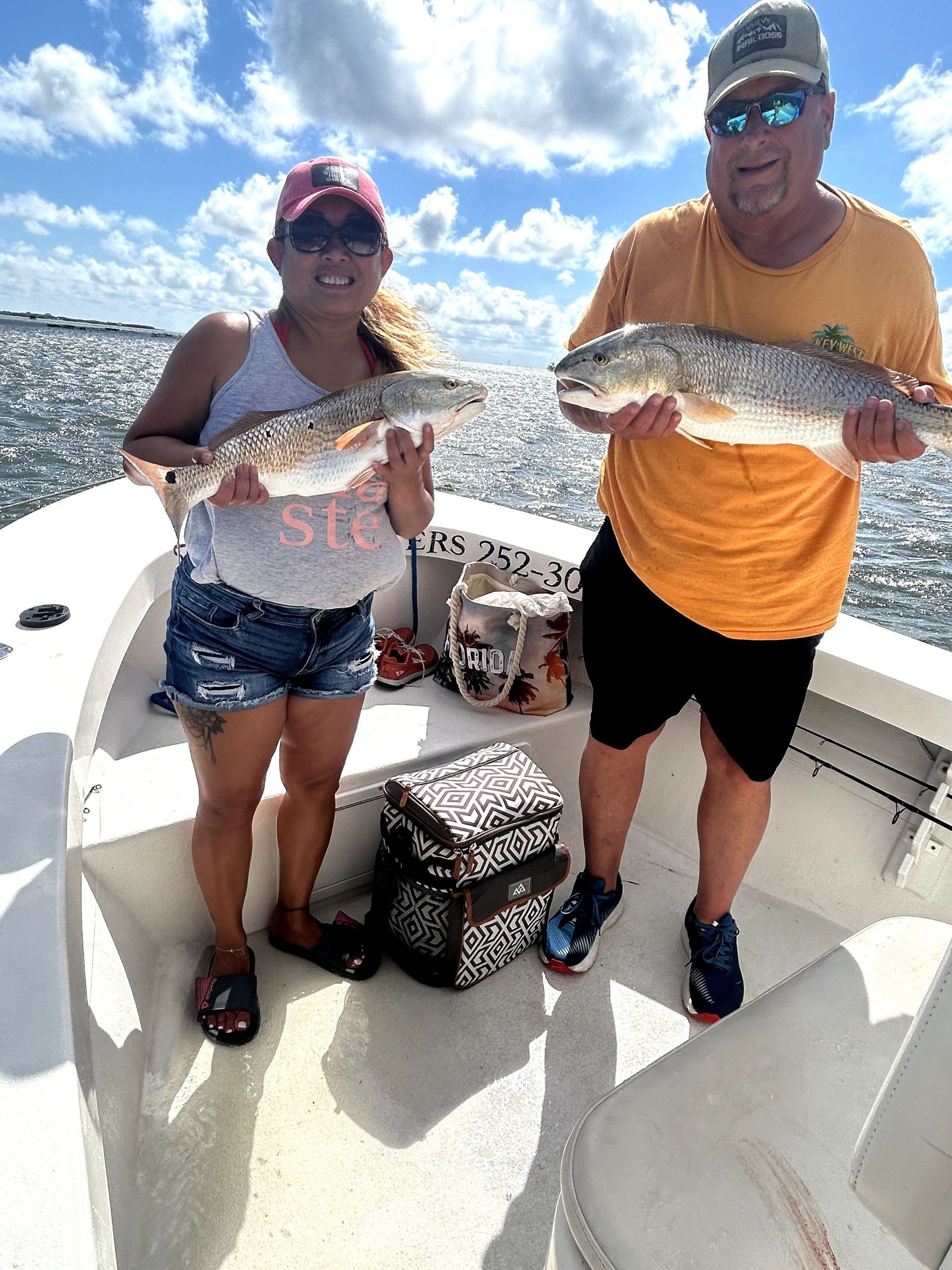 Two anglers on a boat holding large fish under a bright, cloudy sky.