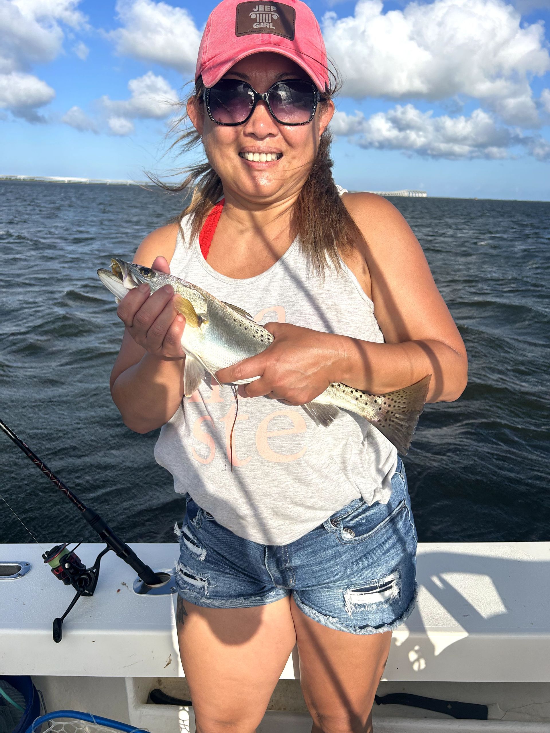 Person smiling and holding a fish on a boat, wearing a pink cap and sunglasses on the water