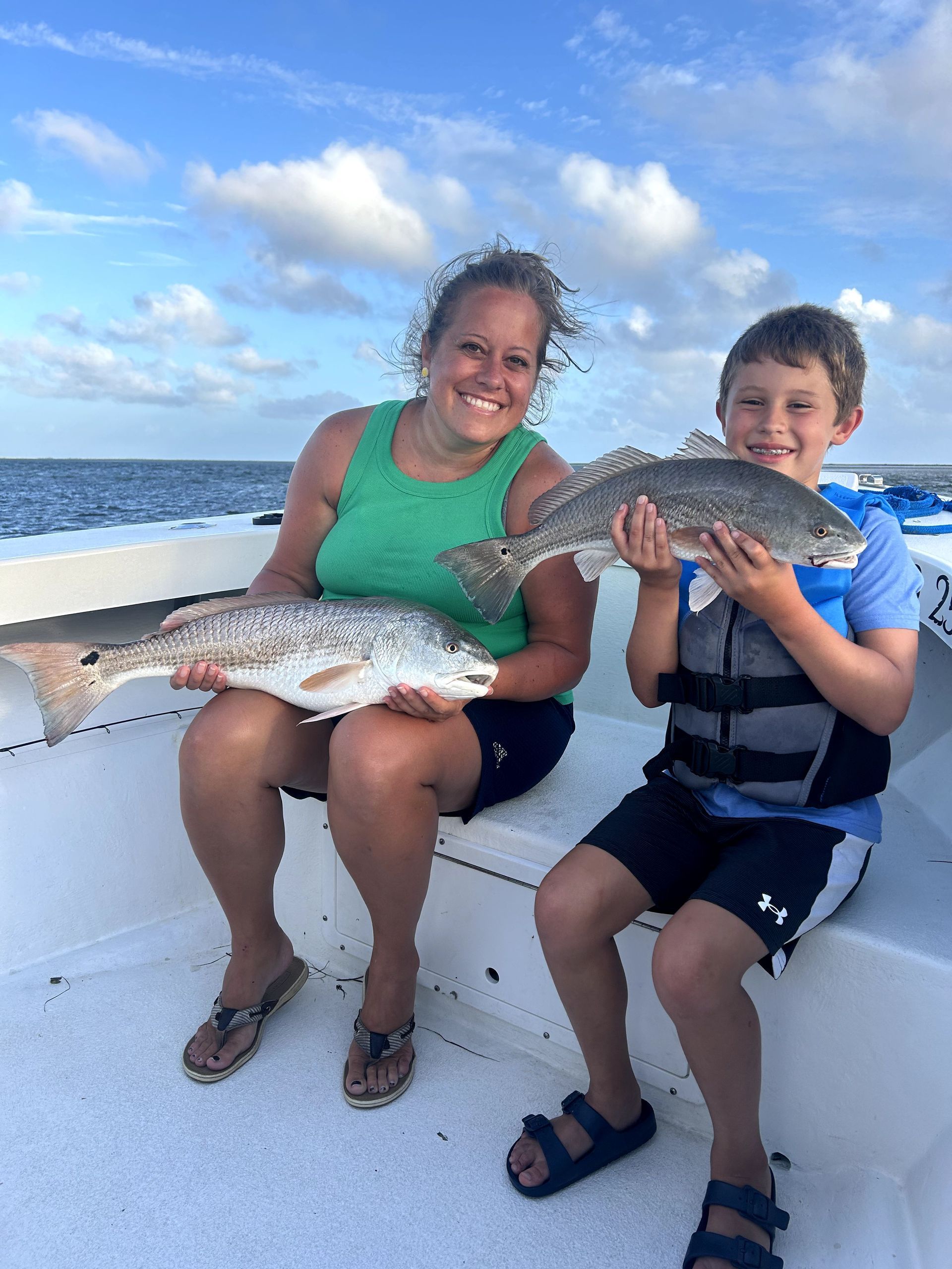 Two children sit on a boat holding large fish, smiling under a blue sky near the water