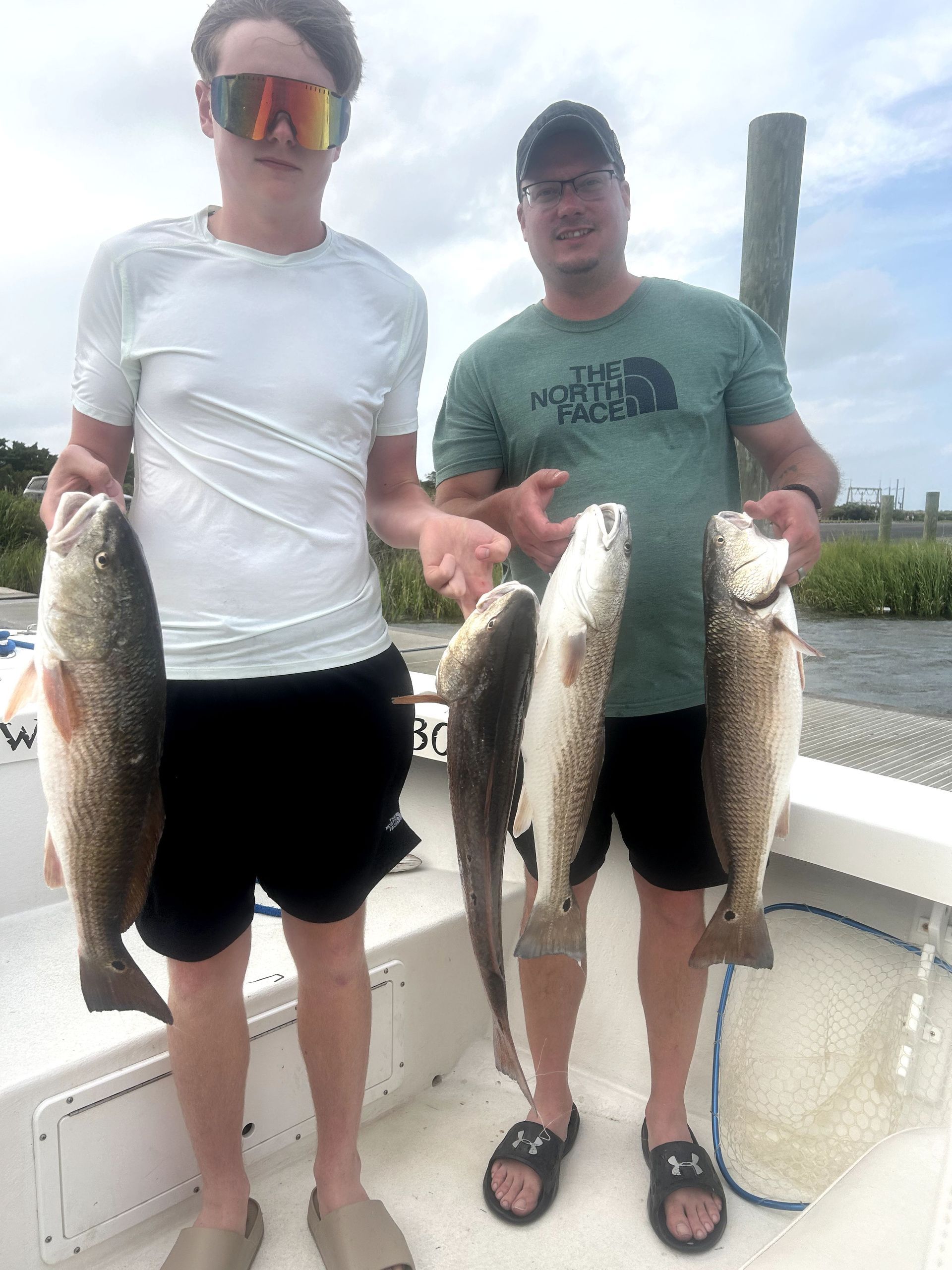 Two people on a boat holding large fish by the water, one in a white shirt and one in a green shirt.