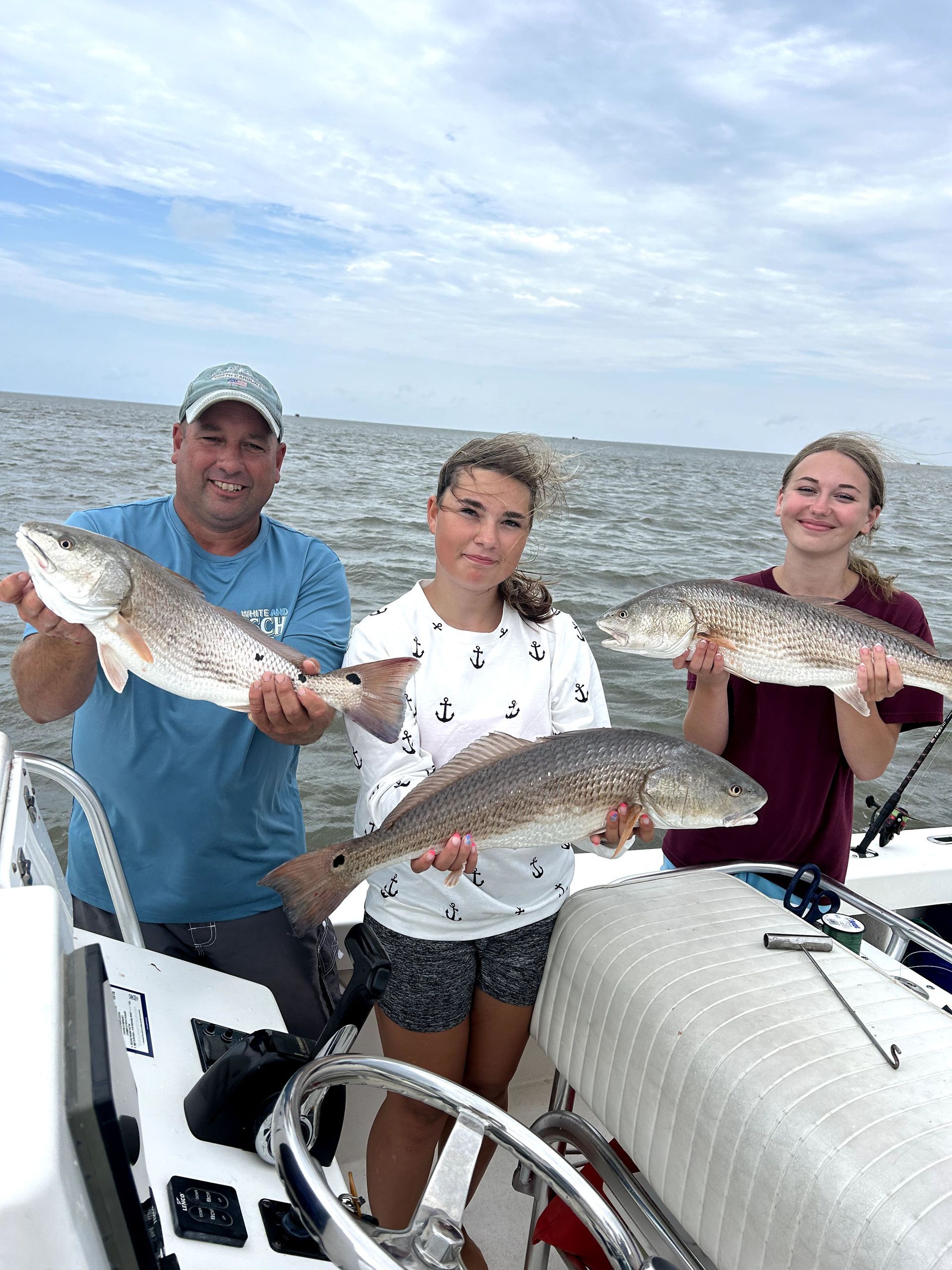 Three people on a boat hold large fish against a cloudy ocean backdrop.