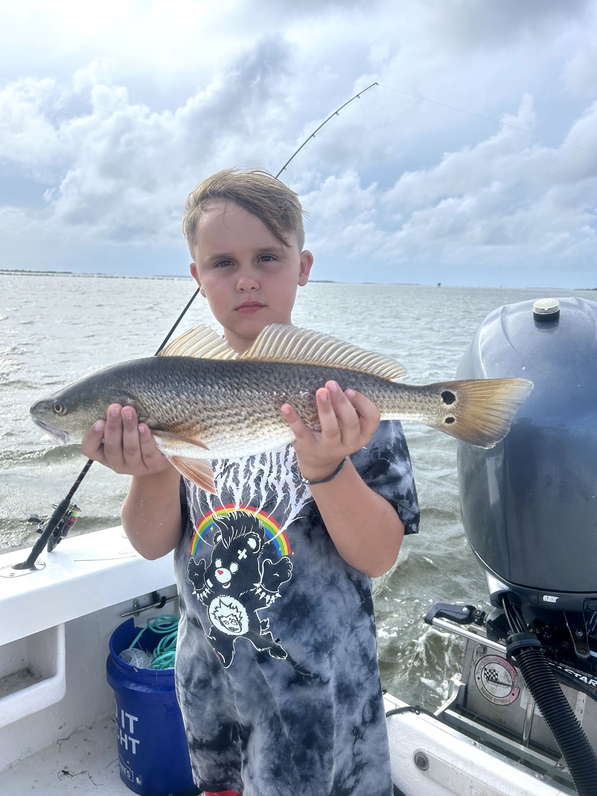 Child holding a large fish on a boat, with a fishing rod and open water behind them.