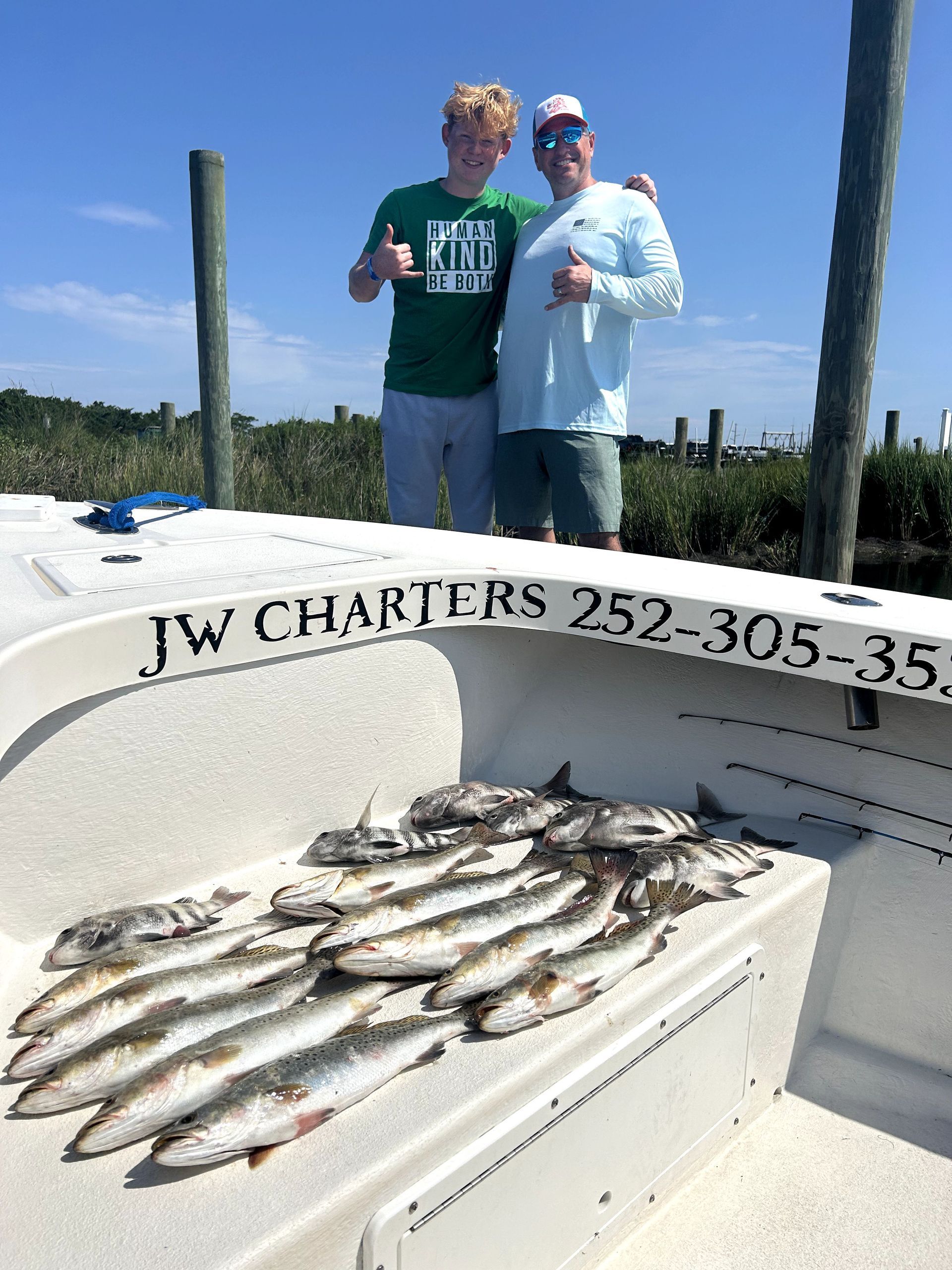 Two people on a boat pose with a large catch of fish on deck under a blue sky.