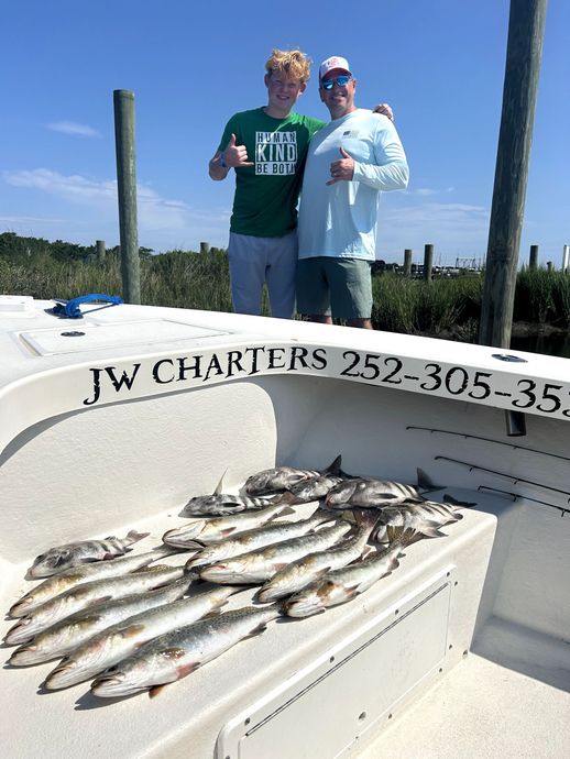 Two people posing on a boat beside a large catch of fish under a bright blue sky