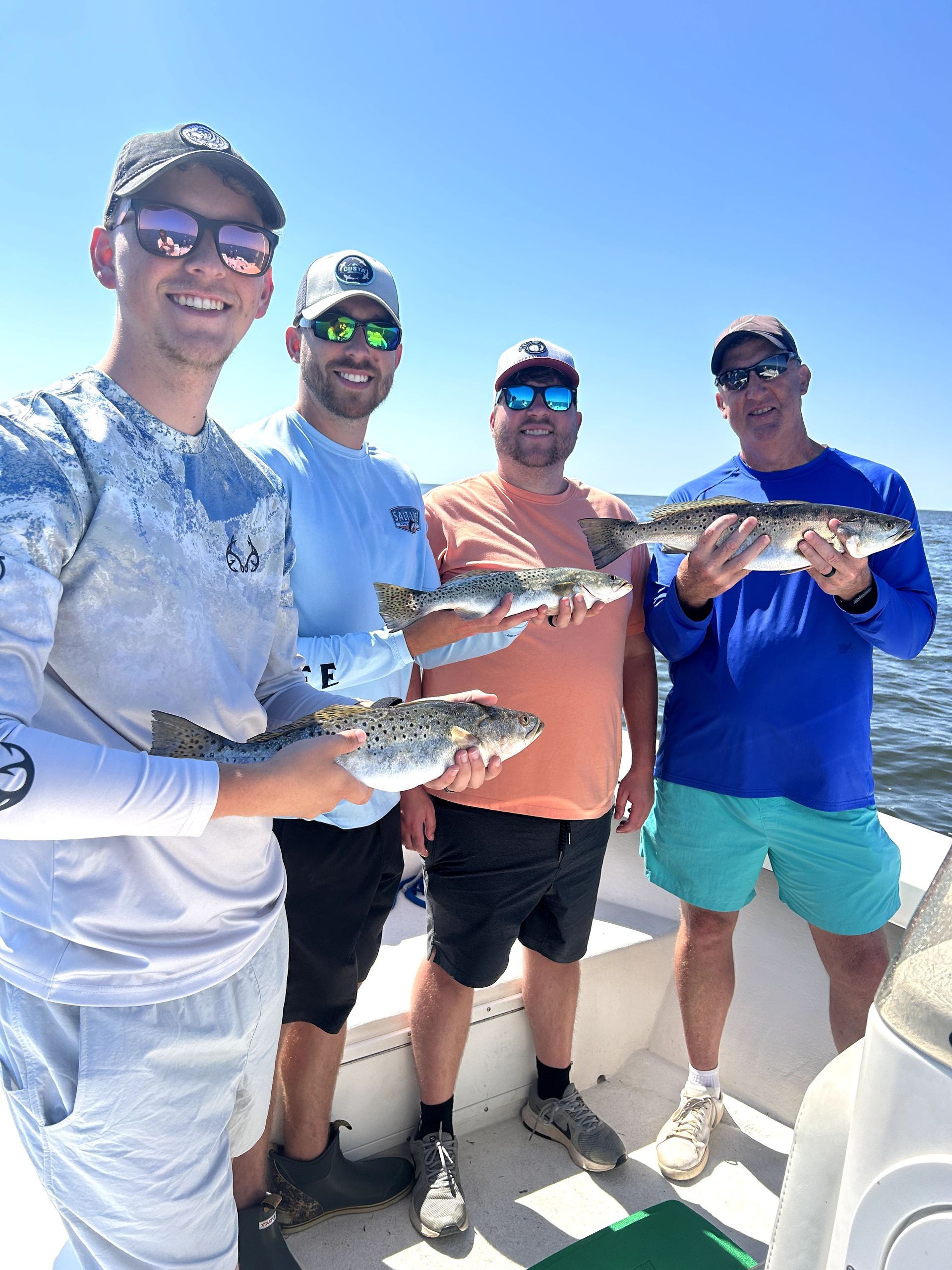 Four people on a boat holding fish, smiling in bright sunlight on open water.