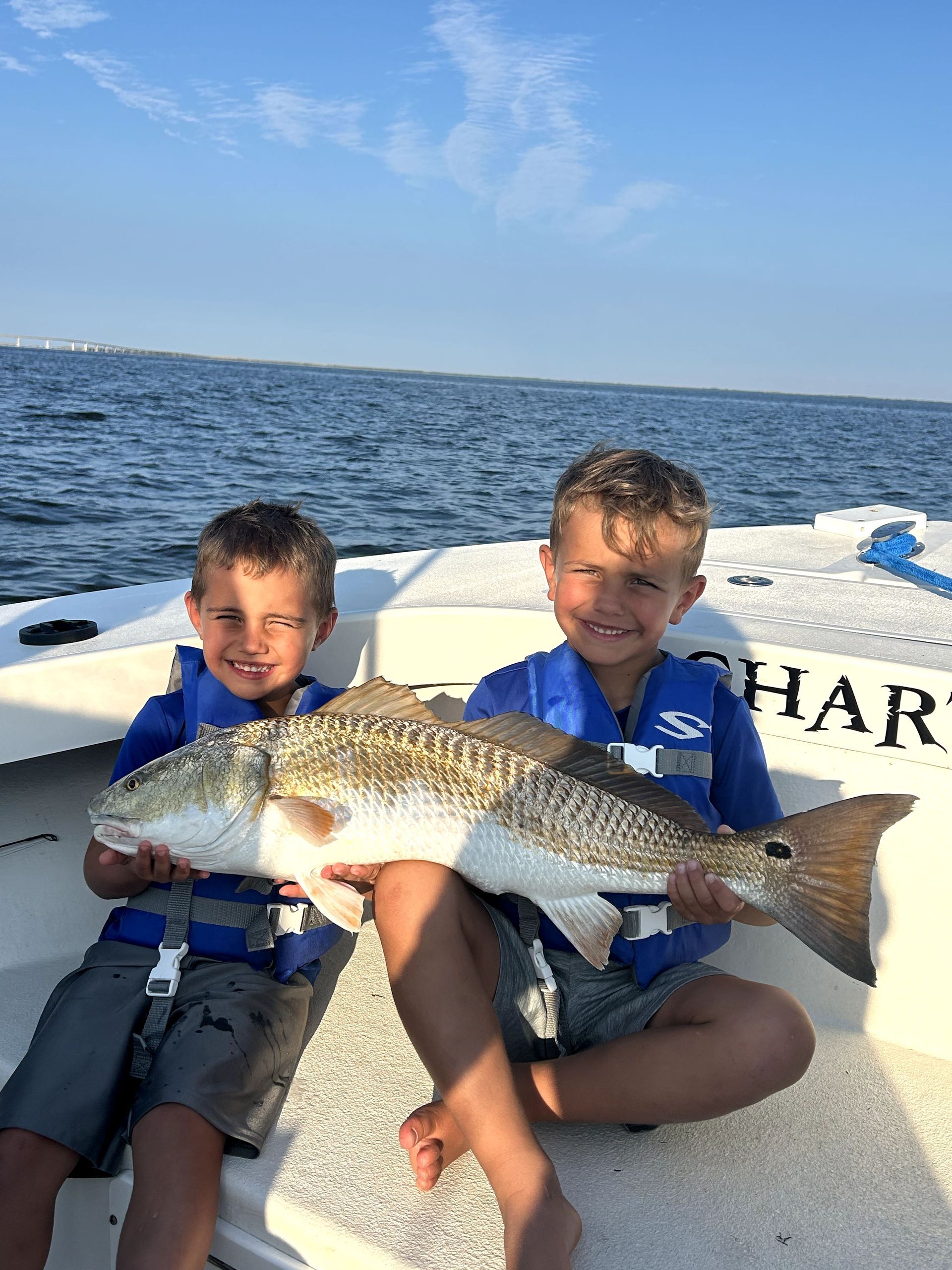 Two children on a boat holding a large fish on the water with blue sky in the background