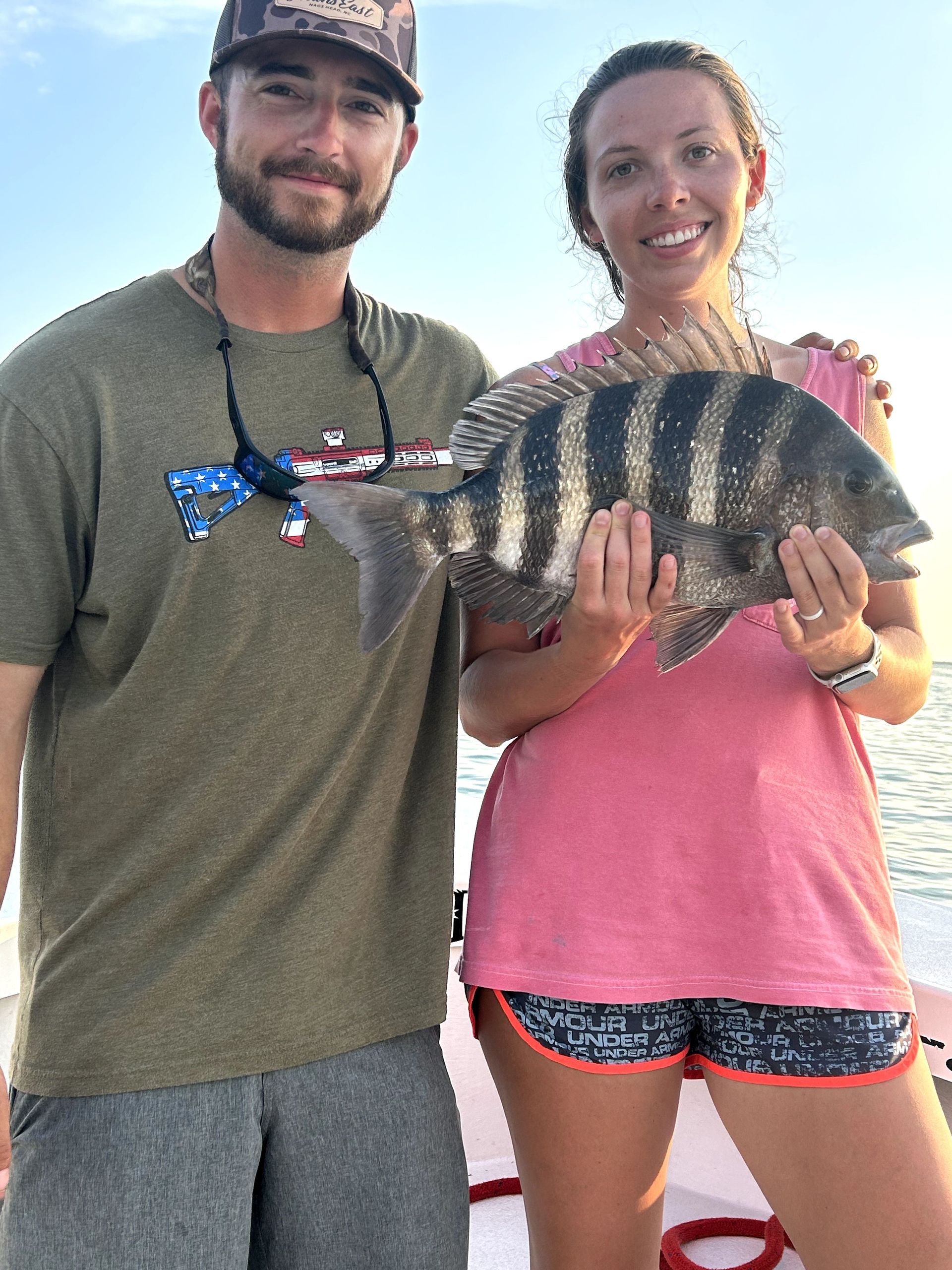 Two people on a boat hold up a striped fish, smiling under a bright sky.