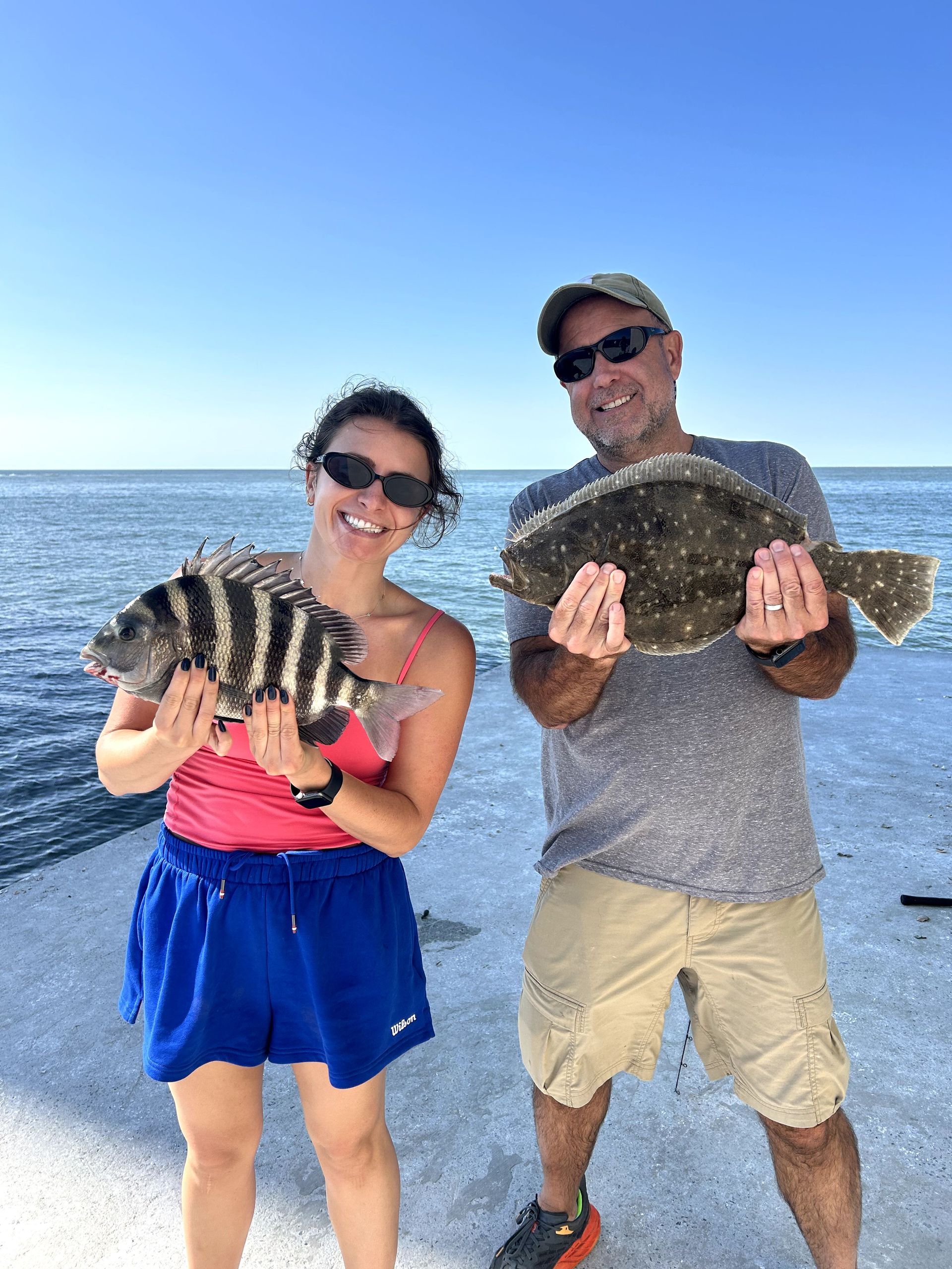 Two people on a beach holding large fish, smiling in bright sunlight.