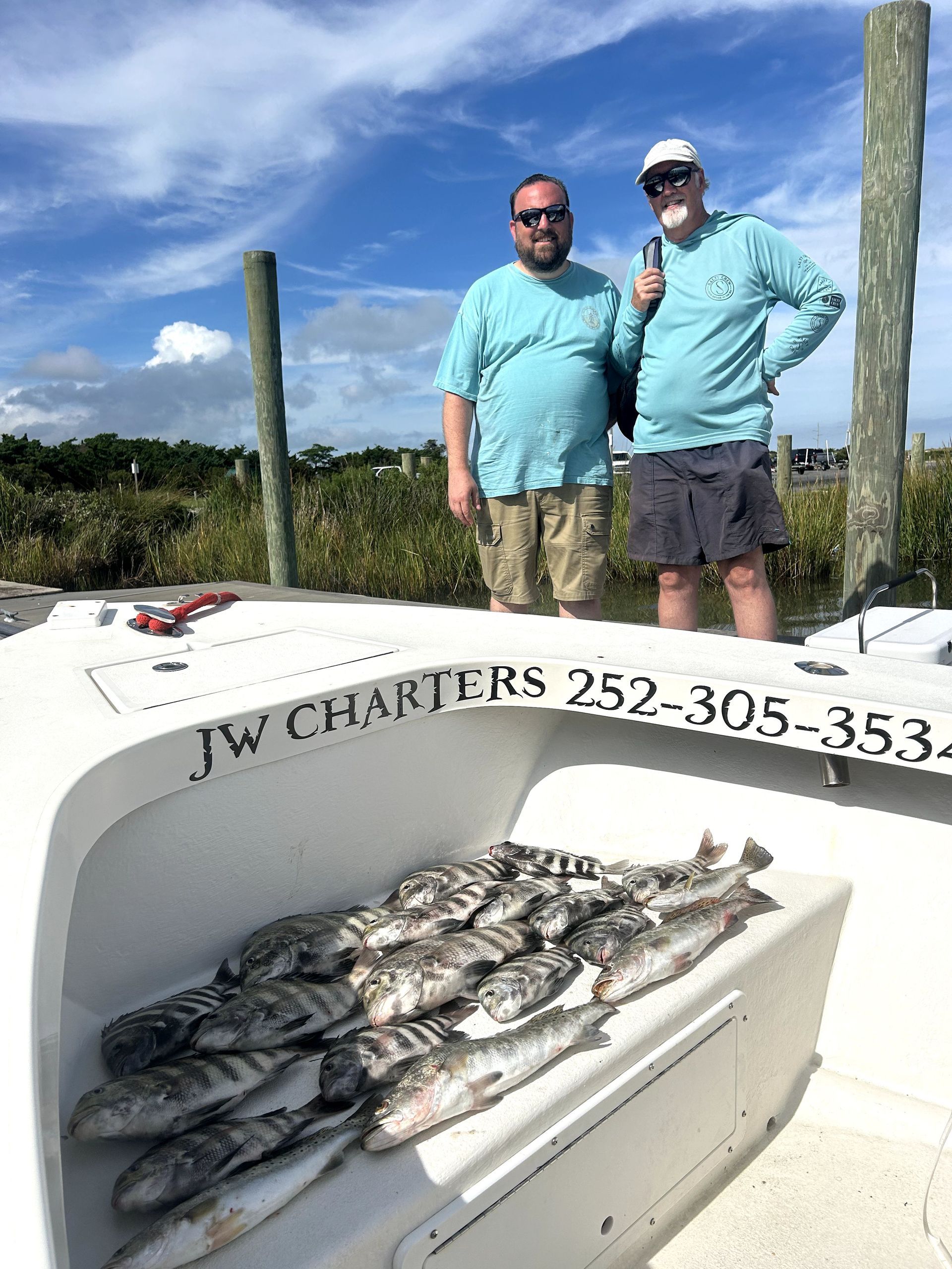 Two men pose on a boat with a pile of fish on deck under a sunny sky.