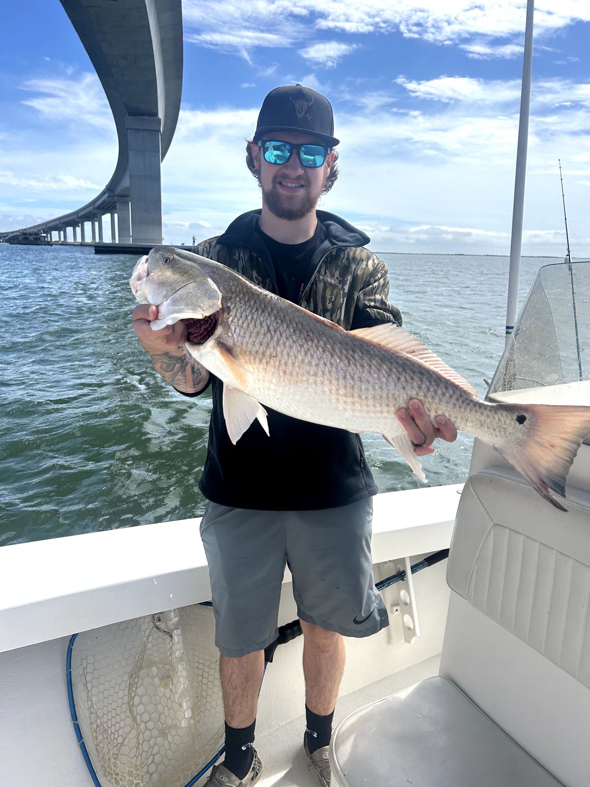 Man holding a large fish on a boat under a bridge, wearing a cap and sunglasses.