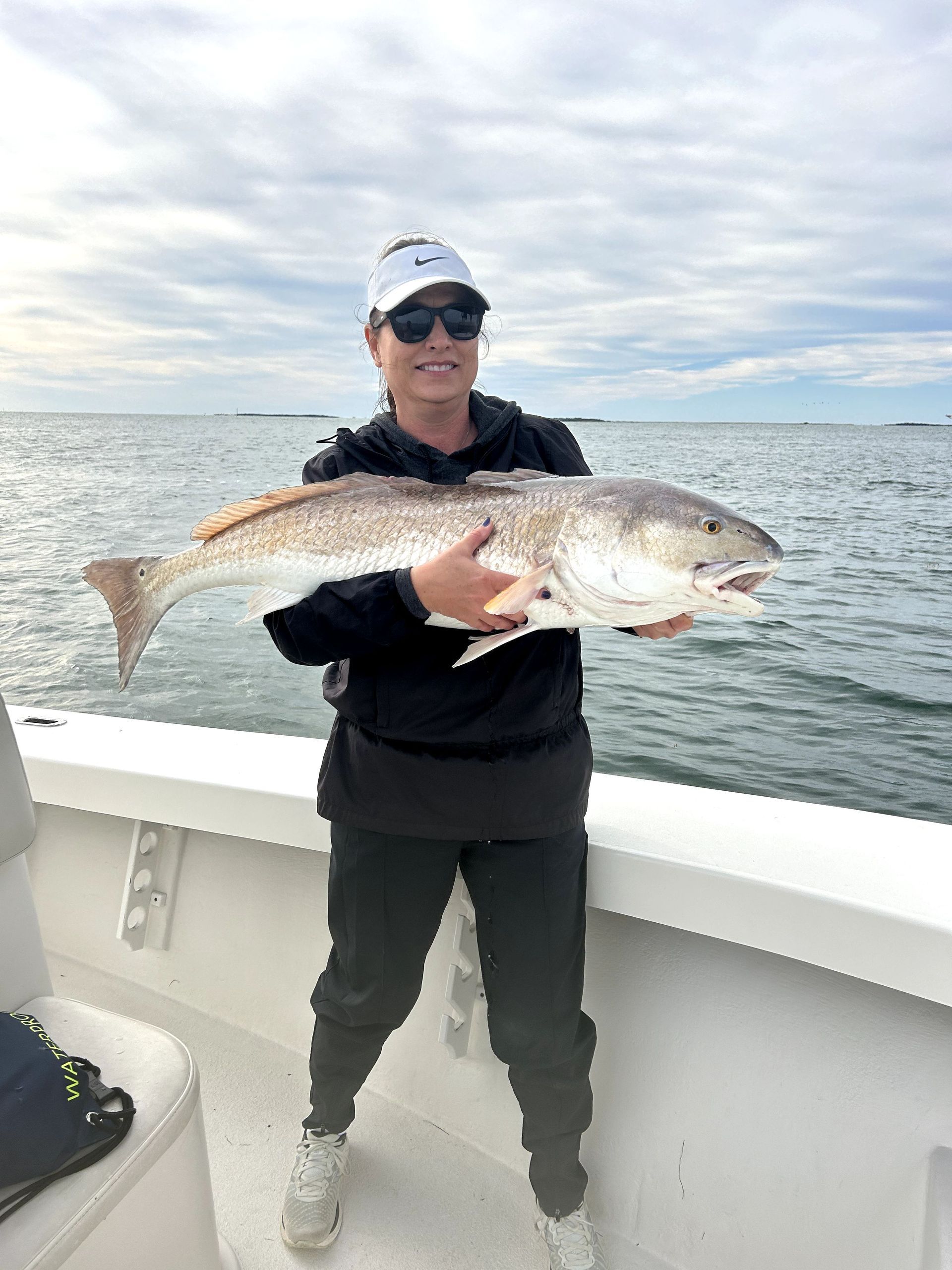 Person on a boat holding a large fish over the ocean, wearing a cap, sunglasses, and dark clothing