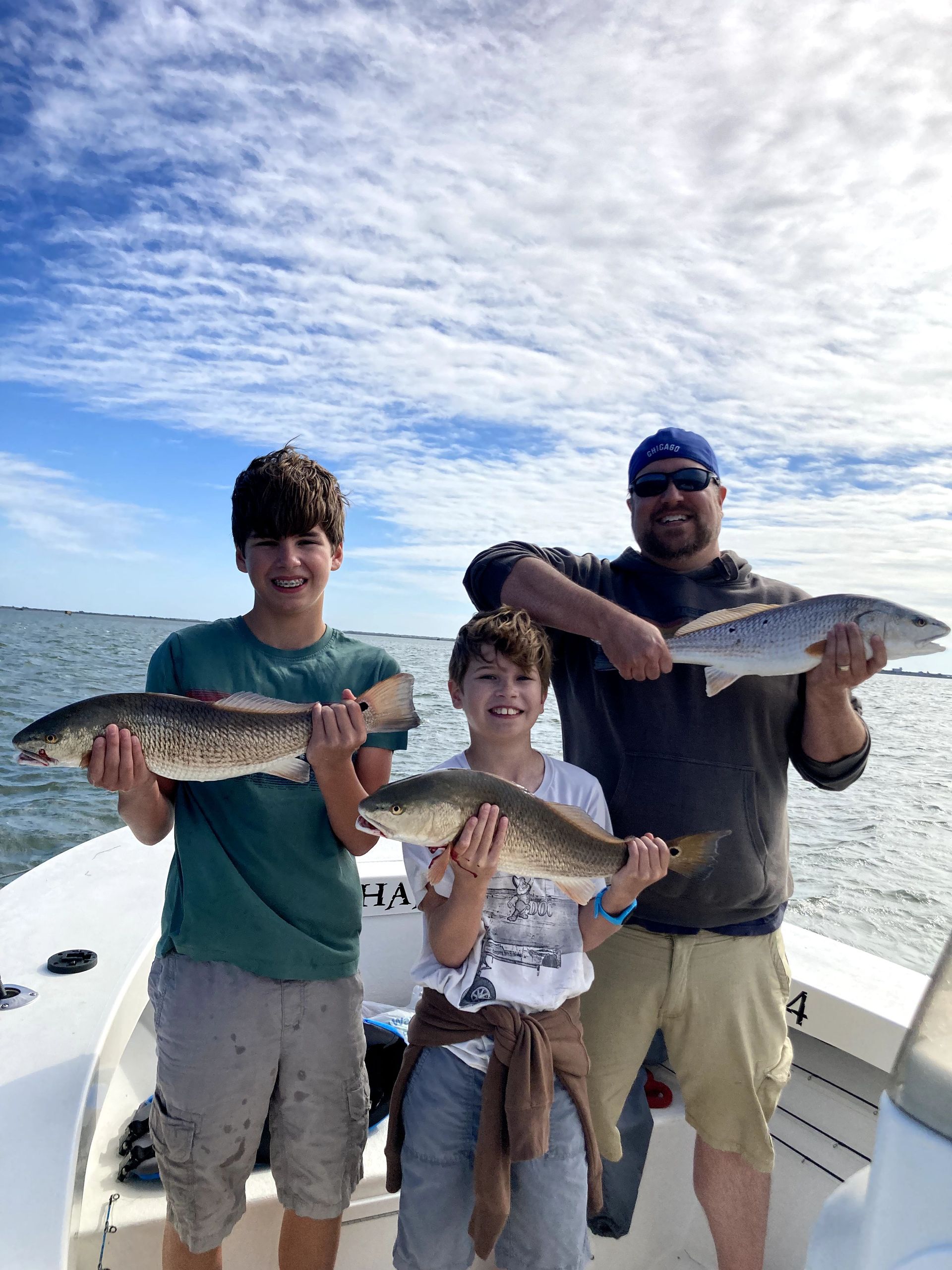 Three people on a boat holding large fish under a cloudy sky.