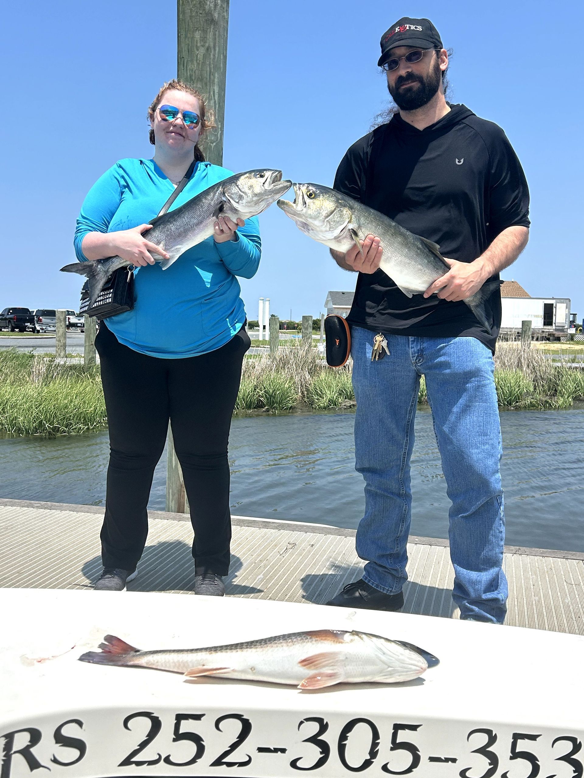 Two people holding large fish outdoors by the water, with another fish laid on a table in front.