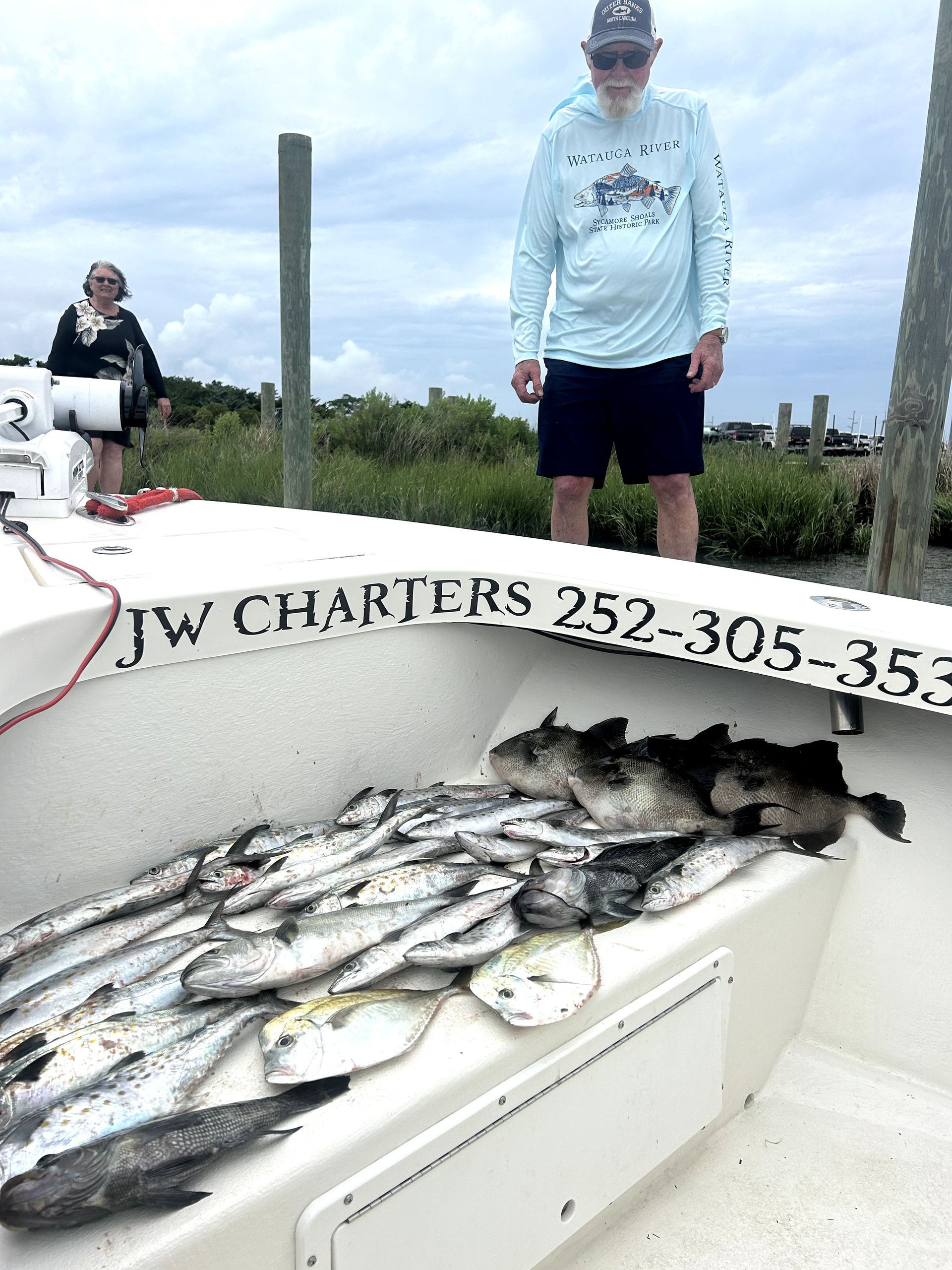 Angler standing on a boat beside a pile of caught fish, with another person in the background.