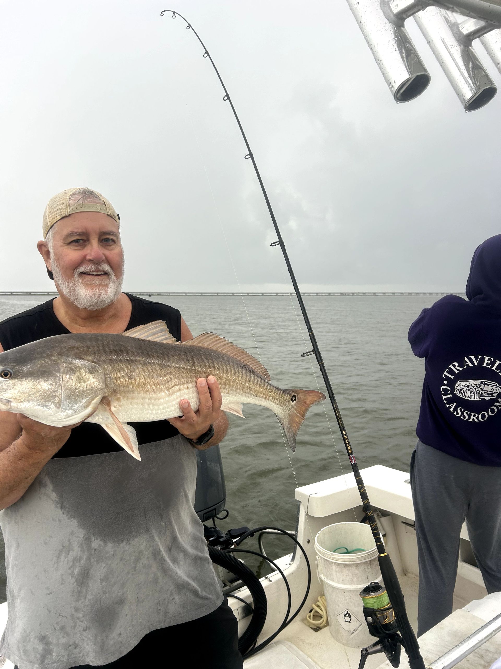 Man holding a large fish on a boat, with another person and fishing gear in the background.