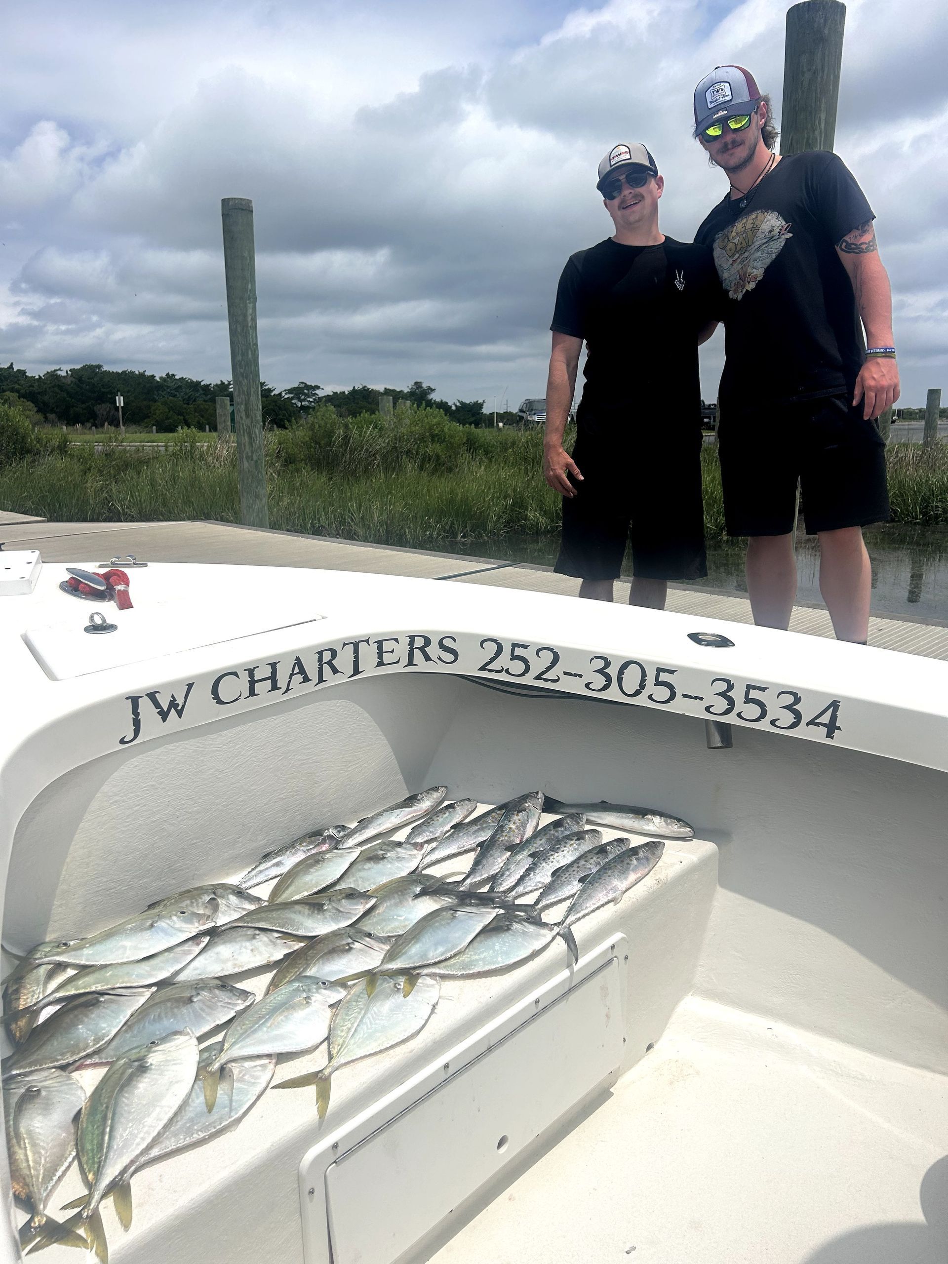 Two people stand on a boat beside a catch of fish on deck under cloudy skies.
