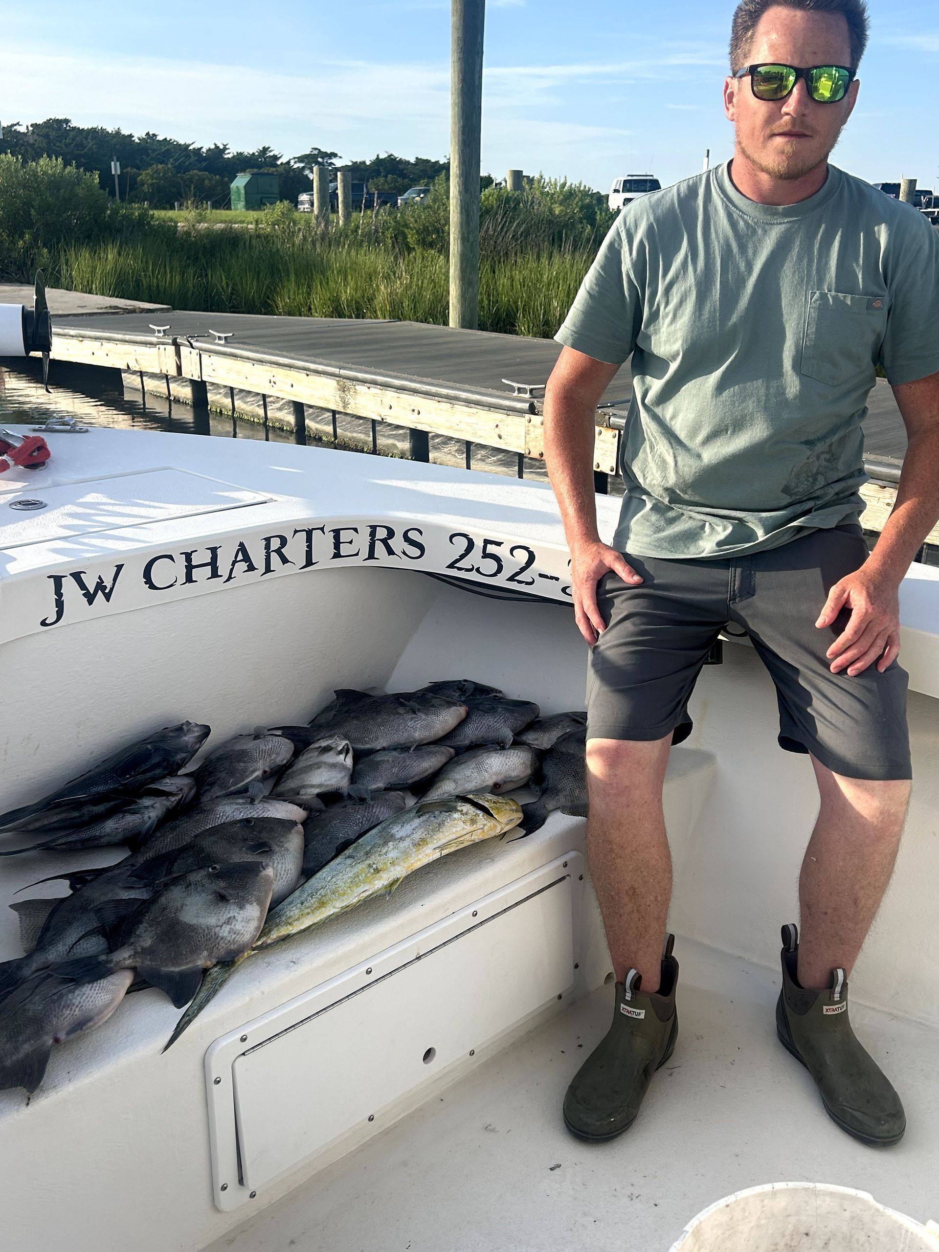 Man standing beside a boat piled with fish on a dock, wearing sunglasses and a gray shirt