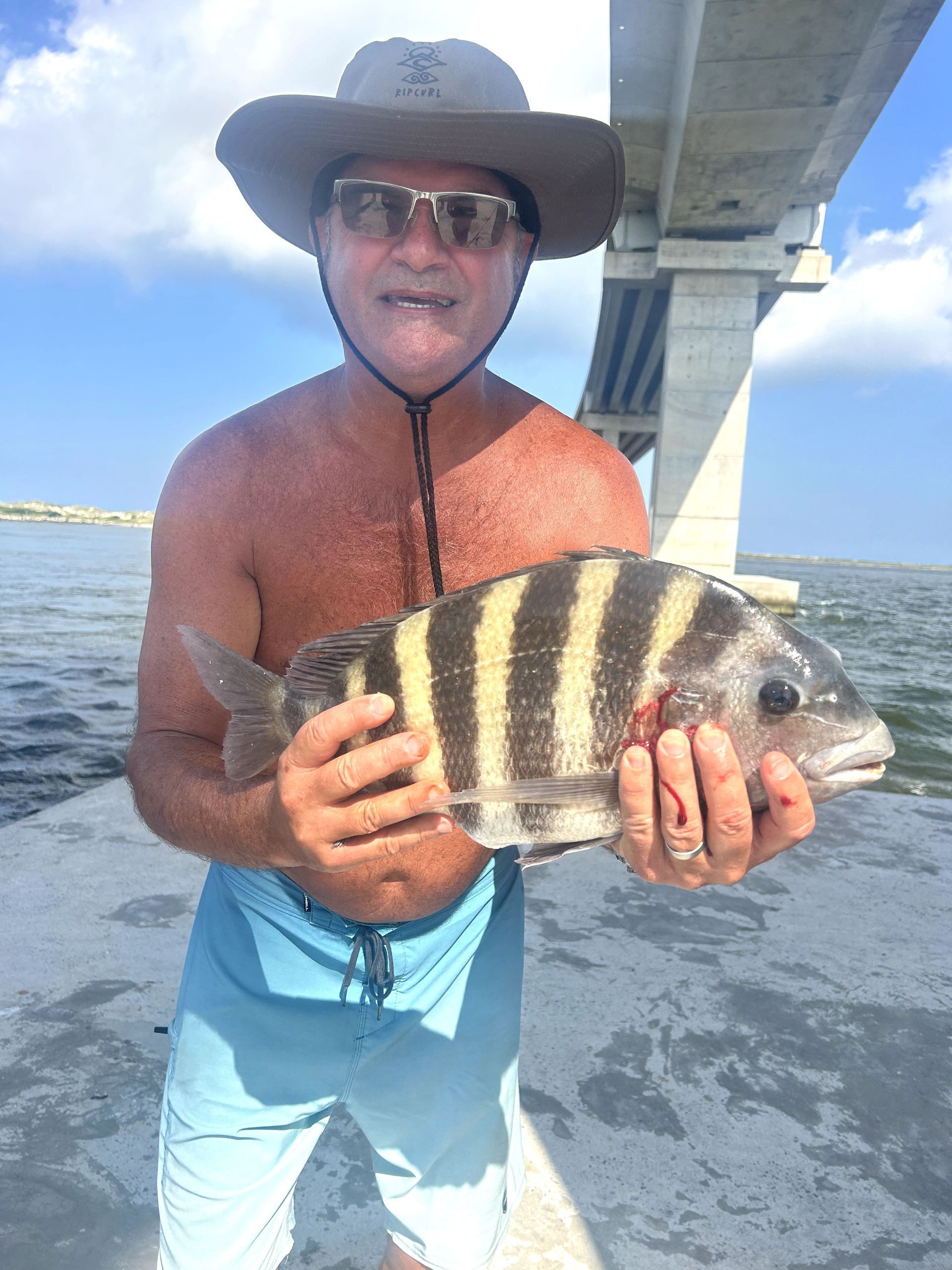 Man holding a striped fish on a pier beside the water, wearing a hat and sunglasses under a bridge.
