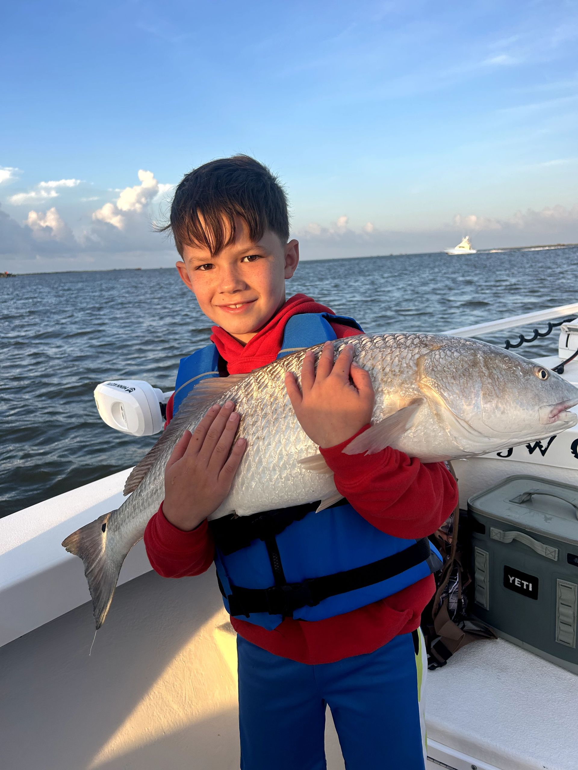 Child holding a large fish on a boat at sea, wearing a red and blue life vest.