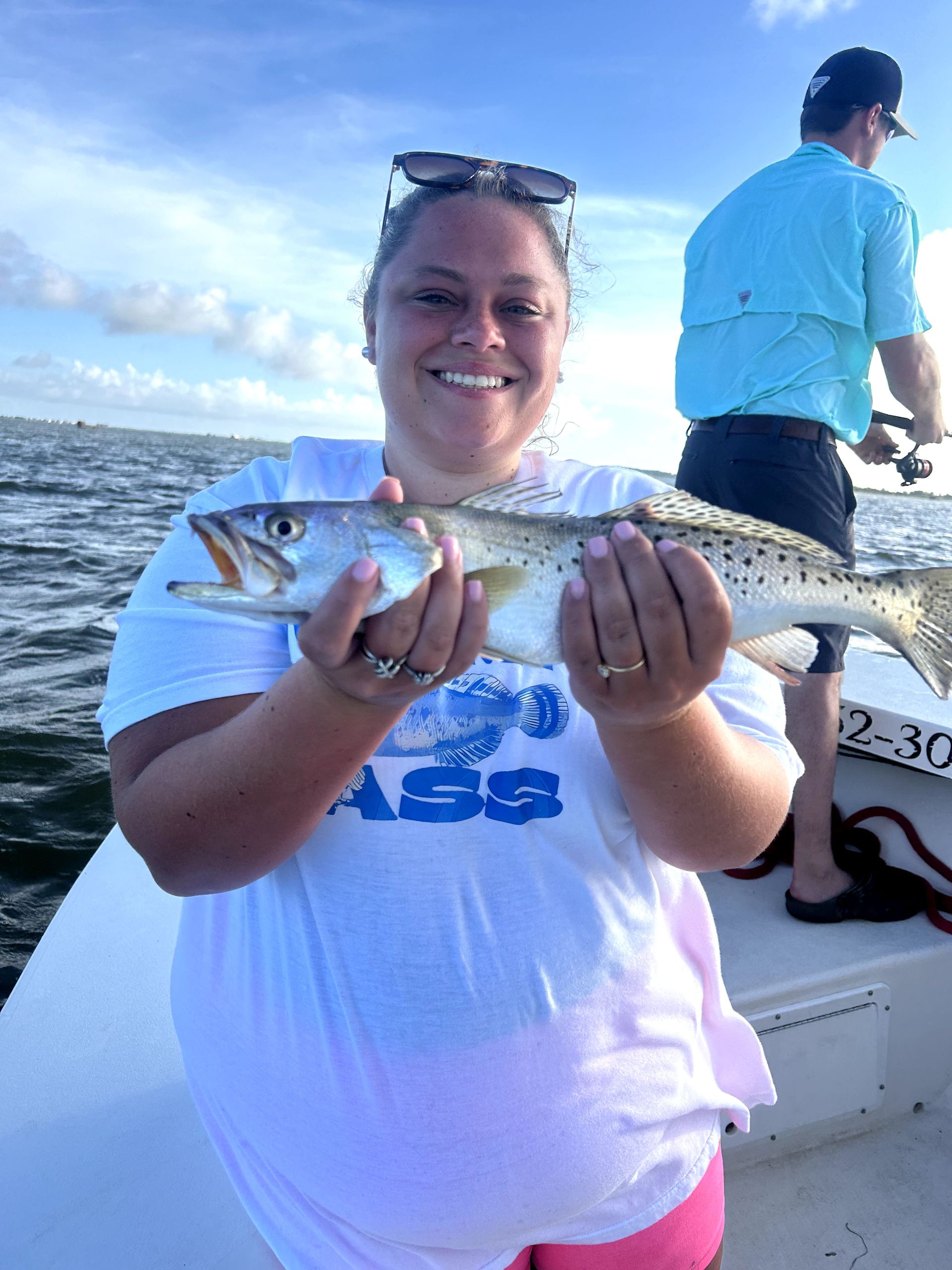 Smiling person on a boat holding a fish, with open water and another person in the background.