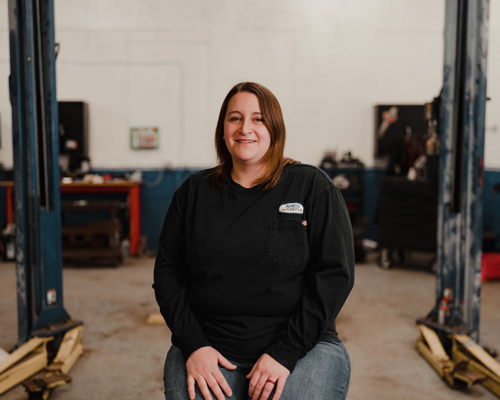 Person sitting in an auto shop between two vehicle lifts, wearing a black sweatshirt and jeans, smiling at camera