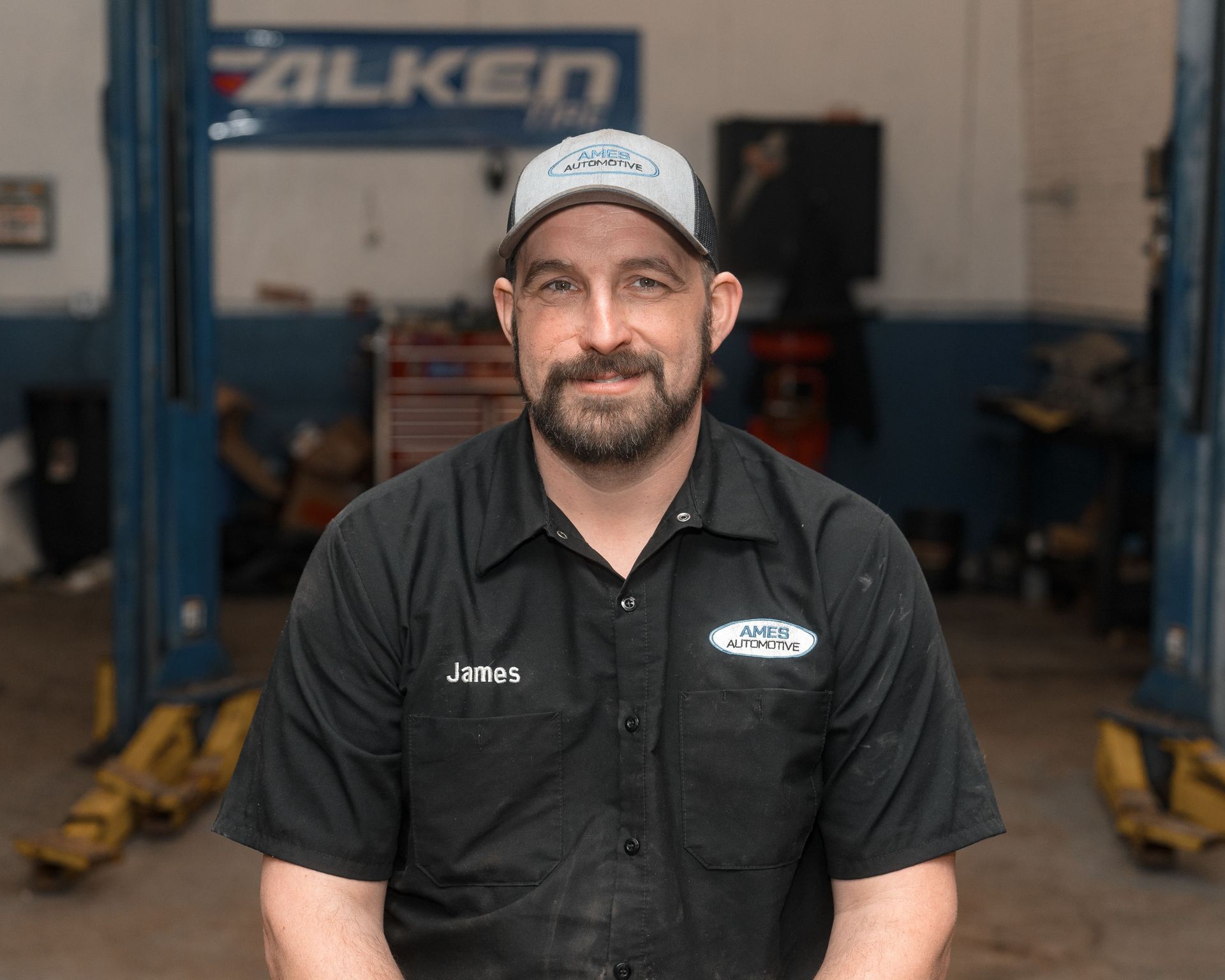 Mechanic in black work shirt and cap seated in an auto shop, with lifts and tools in the background