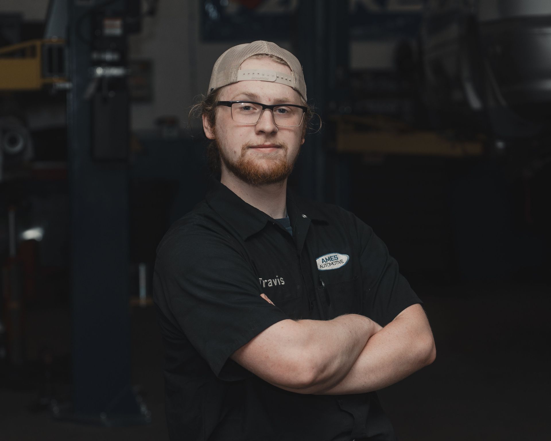 Person in a black work shirt and cap stands with arms crossed in a dim industrial setting.