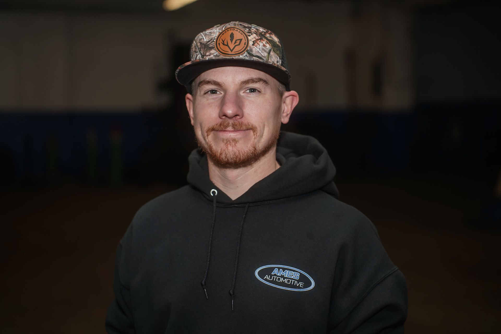 Smiling man in a black hoodie and patterned cap against a dark indoor background