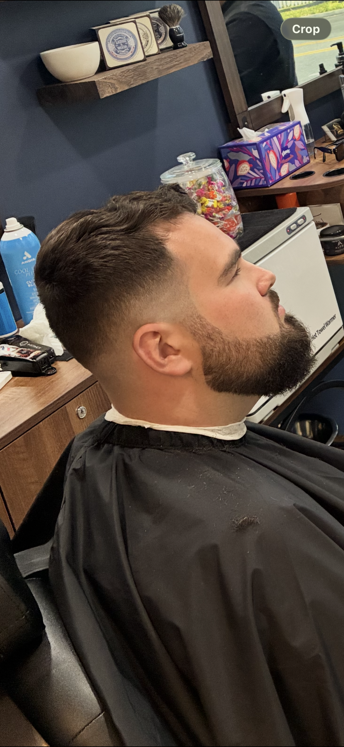 Man with a neat beard getting a haircut in a barbershop, shown in side profile.