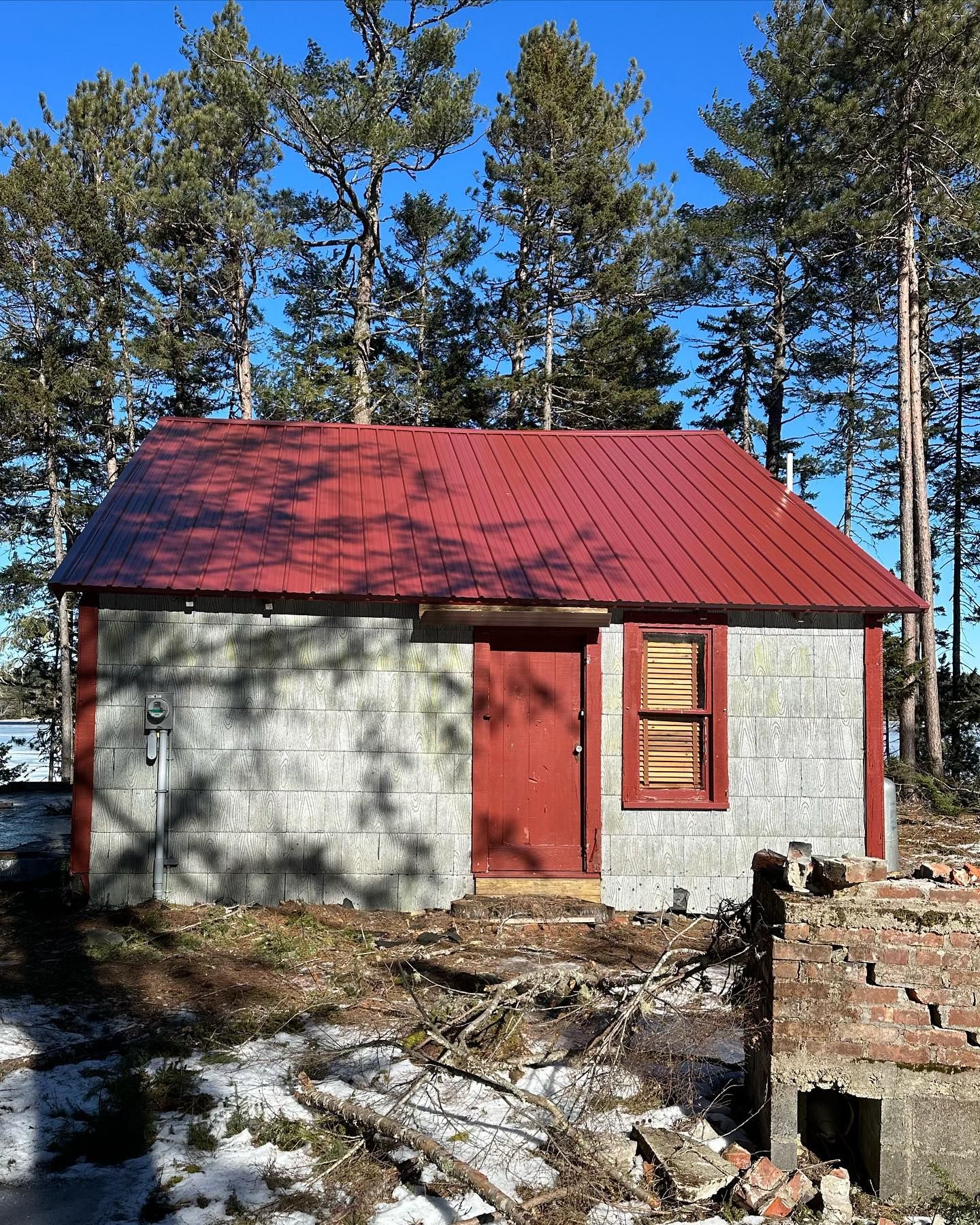 Small gray building with a red door and roof, surrounded by trees on a sunny day.