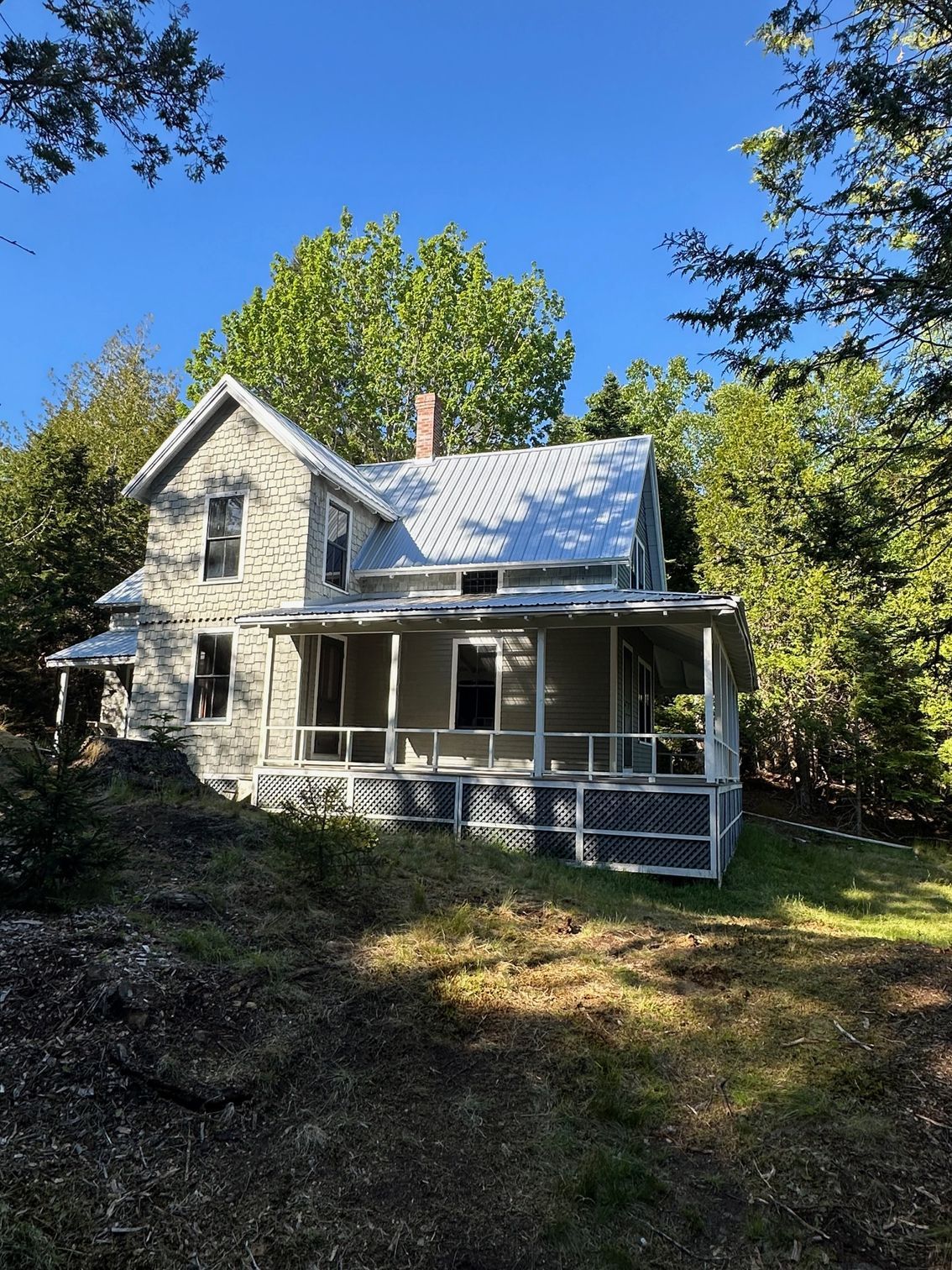 Old stone house with porch and screened-in area, surrounded by trees under a blue sky.