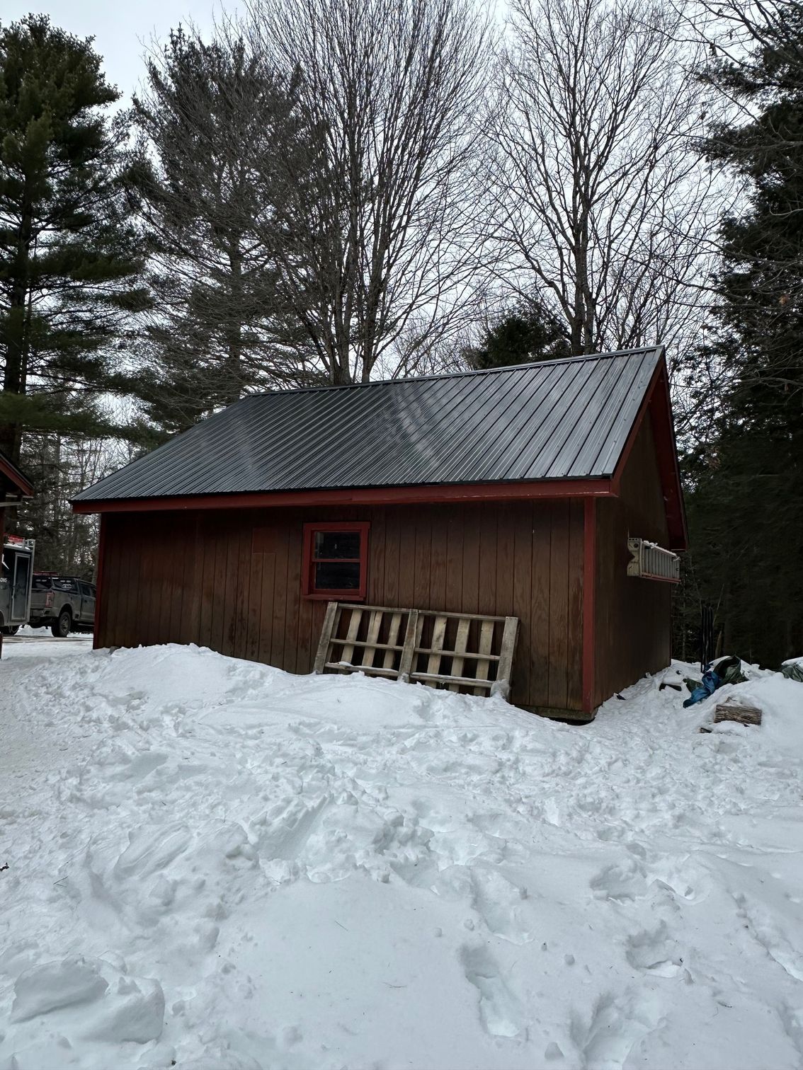 Brown shed with a metal roof in a snowy area. Pallets and a small window are visible.