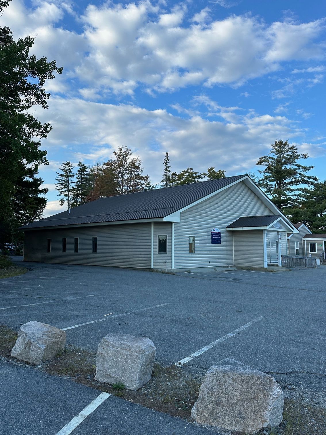 Gray building with a dark roof and a small covered entrance. Parking lot in front, cloudy sky.