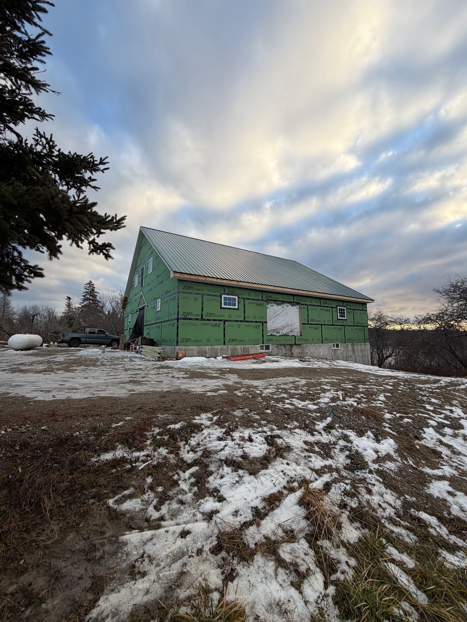 A two-story house under construction with green siding and a partially covered roof, in a snowy field under a cloudy sky.