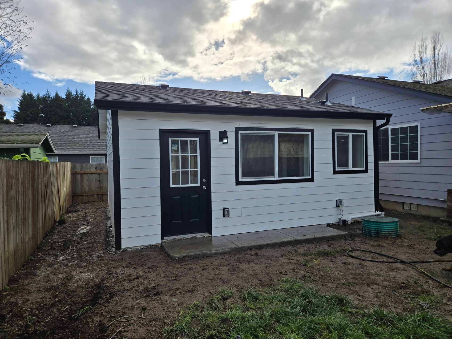 Small white backyard shed with black door and window, shown in a dirt yard under a cloudy sky