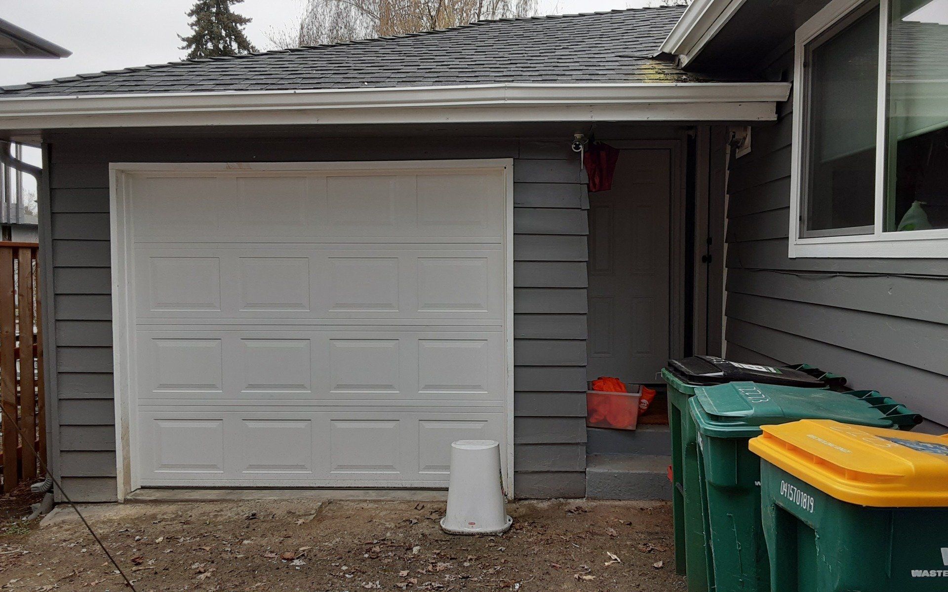 Garage with white door beside gray house, with green trash bins and a yellow-lidded bin in front.