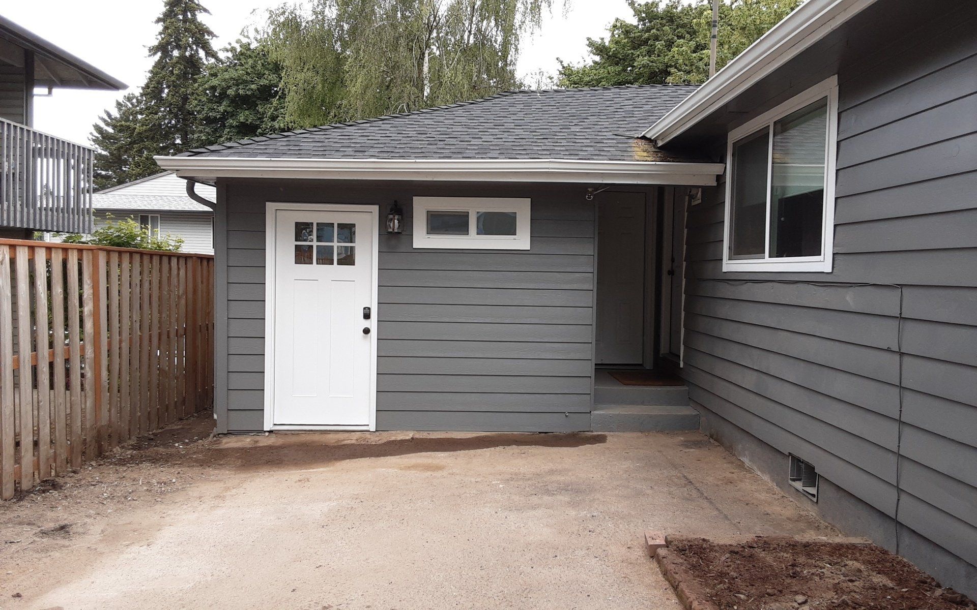 Small gray backyard shed with white door and window beside a house, on a concrete patio.