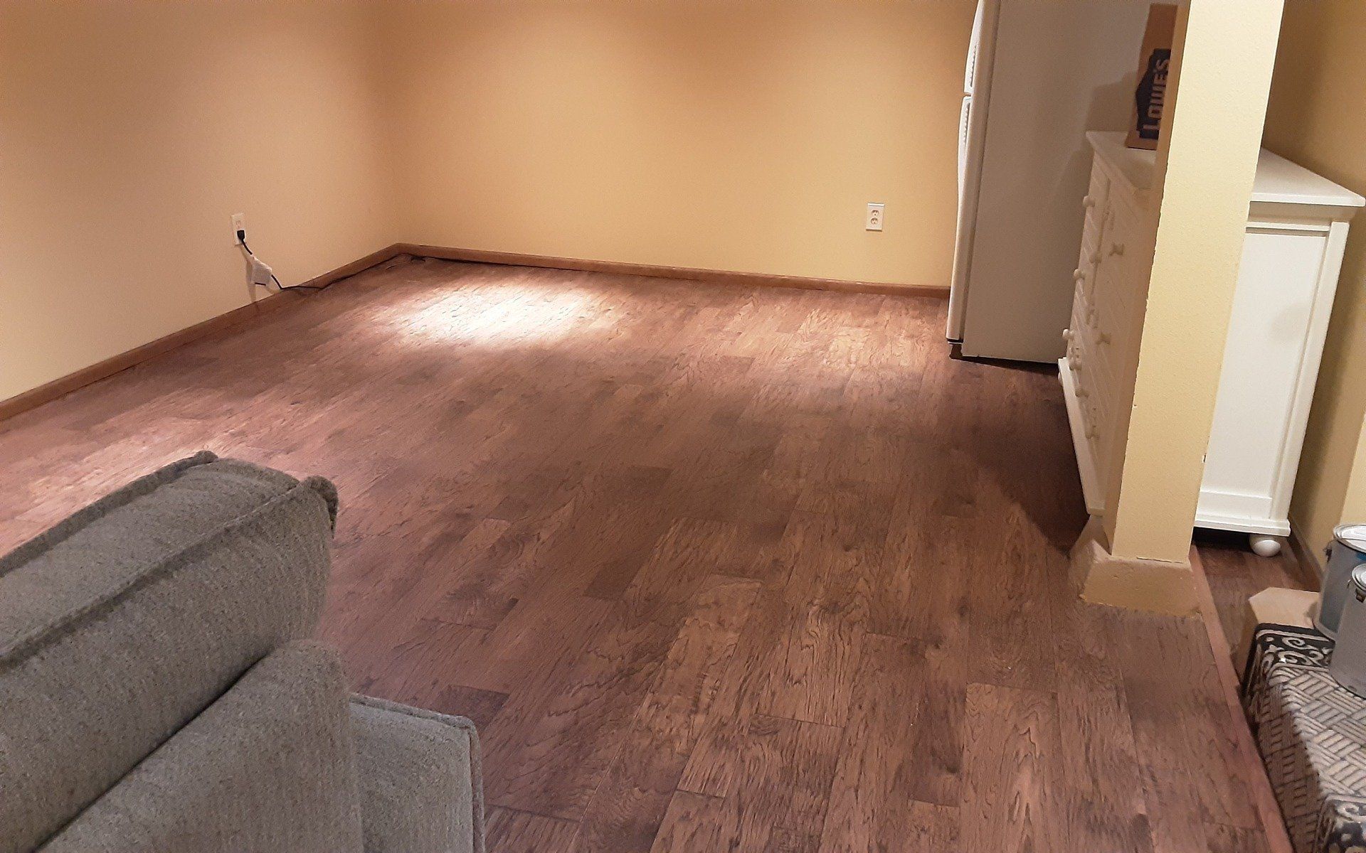 Empty carpeted living room with beige walls, gray sofa corner, and doorway to a kitchen area.