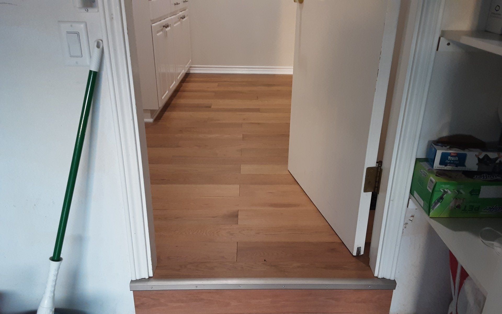 Hallway with wood flooring and a green broom leaning against a white wall near an open door.