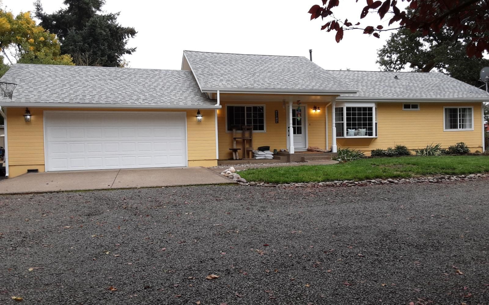 Yellow ranch-style house with white garage door, covered porch, and gravel driveway under overcast sky