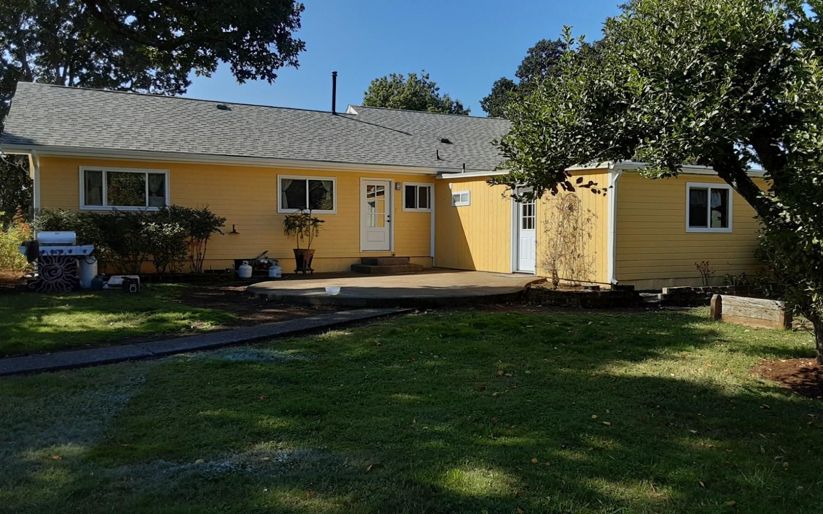 Yellow single-story house with white trim, front door, and lawn under trees on a sunny day