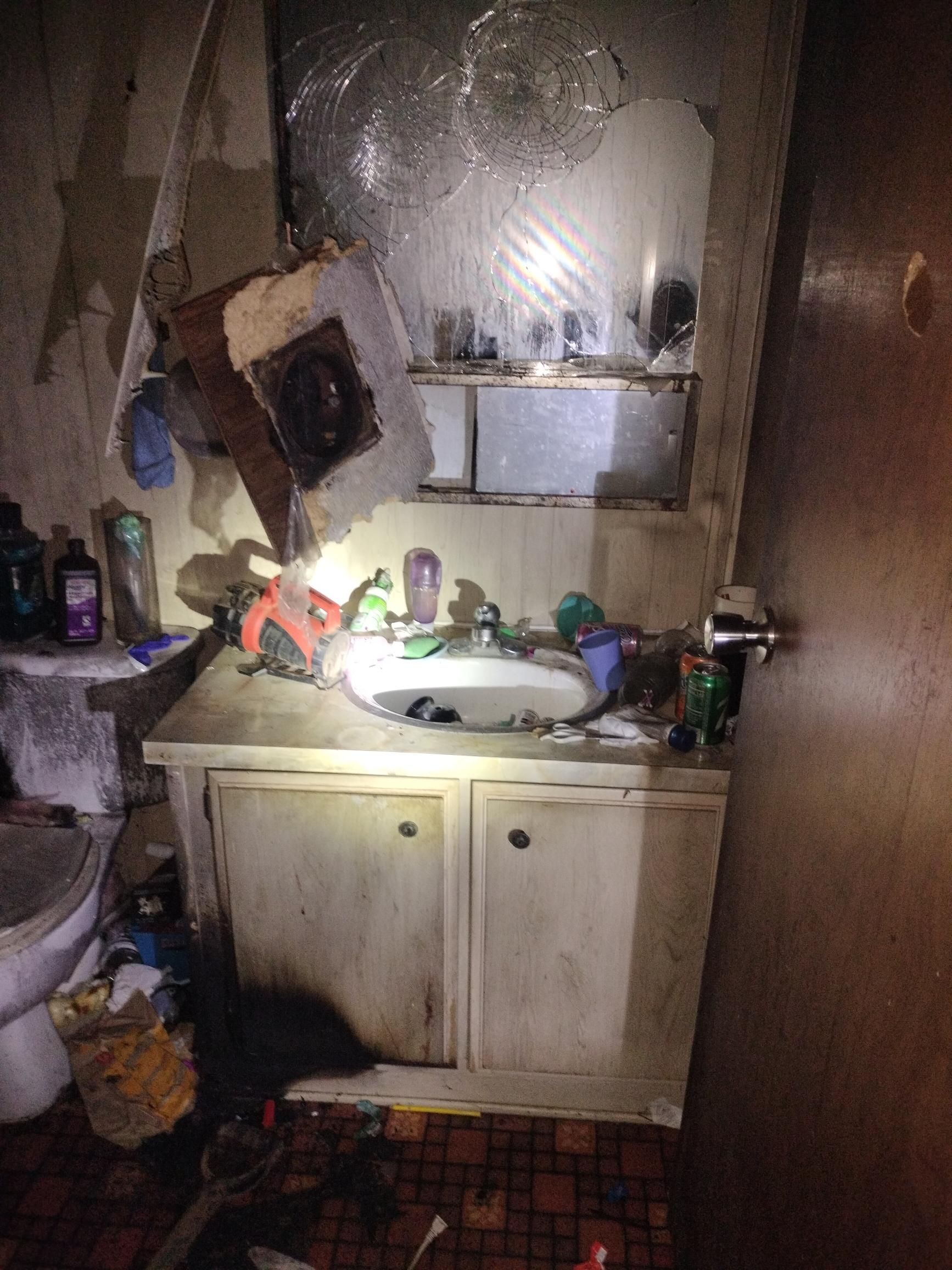 Cluttered bathroom with a stained sink cabinet, mirror, and toiletries piled around the sink.