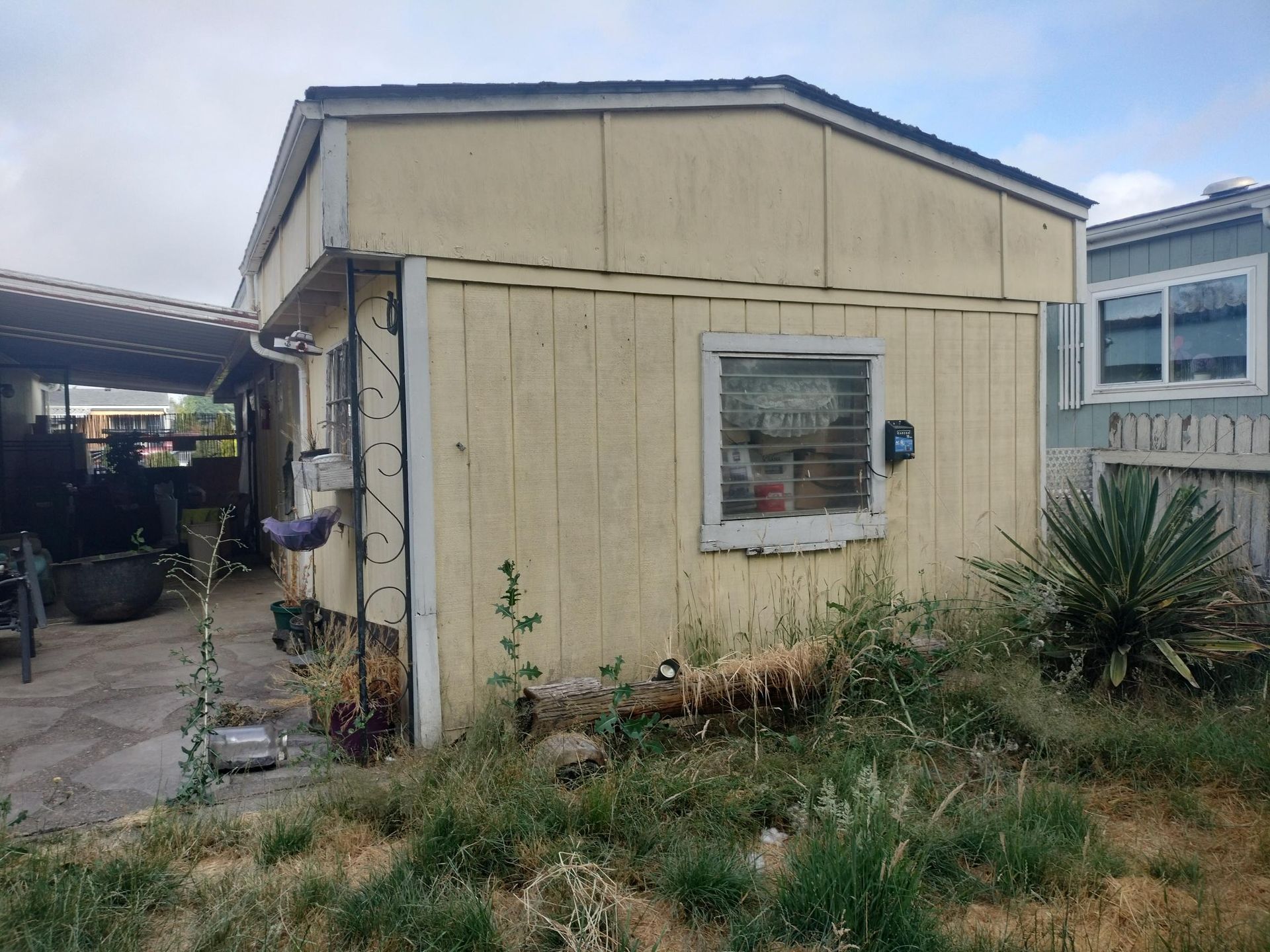 Small beige shed with a window in a grassy yard beside a covered carport.