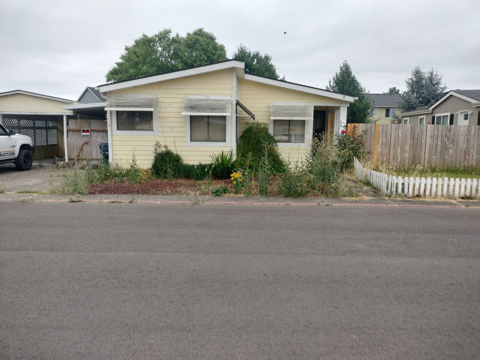 Small beige house with overgrown front yard and white fence beside a street