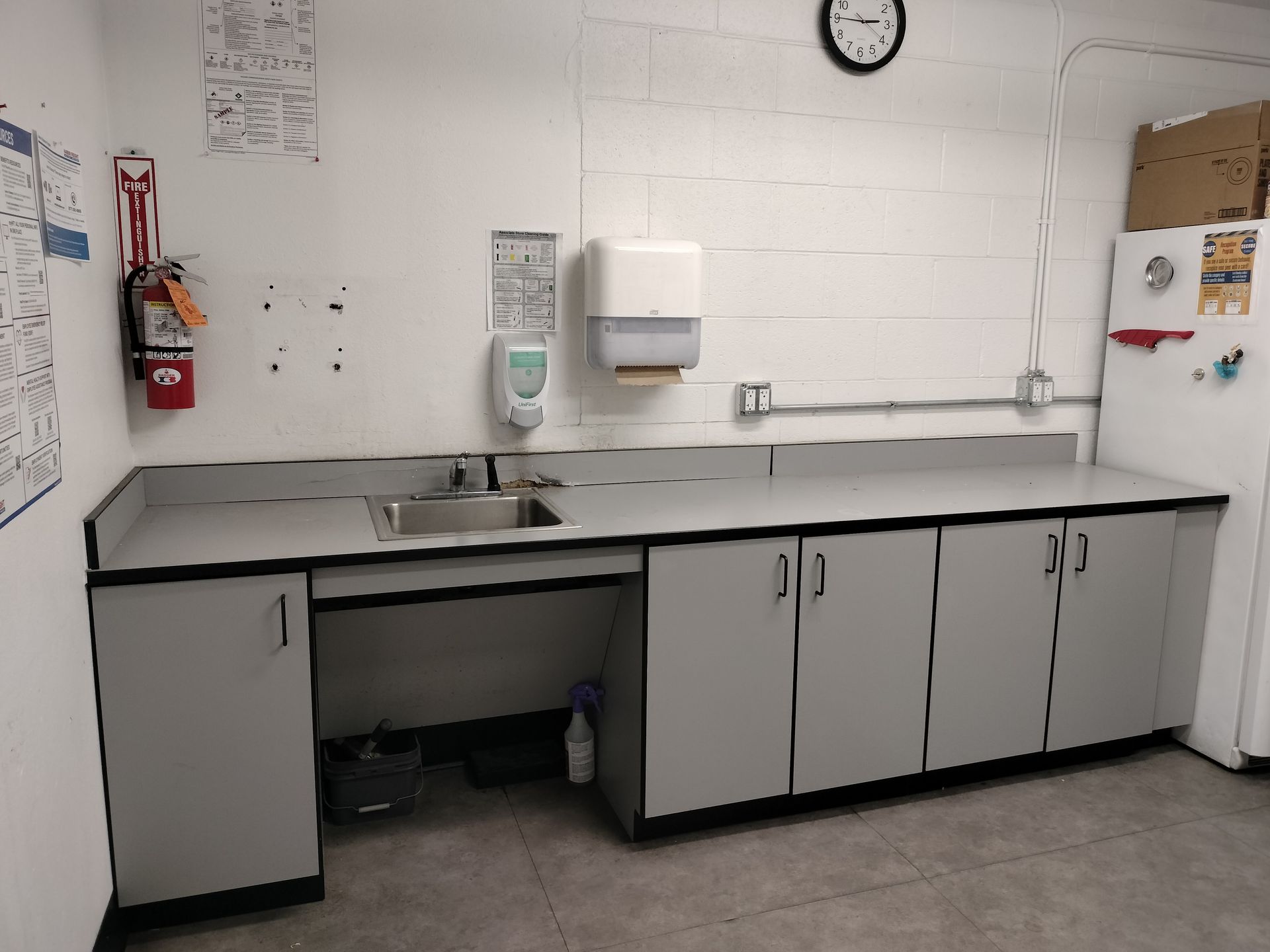 Empty breakroom sink and counter with cabinets, soap dispenser, wall clock, and fire extinguisher