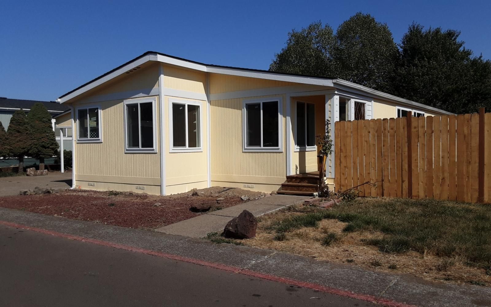 Small beige house with white trim and a wooden fence beside a road under a clear blue sky