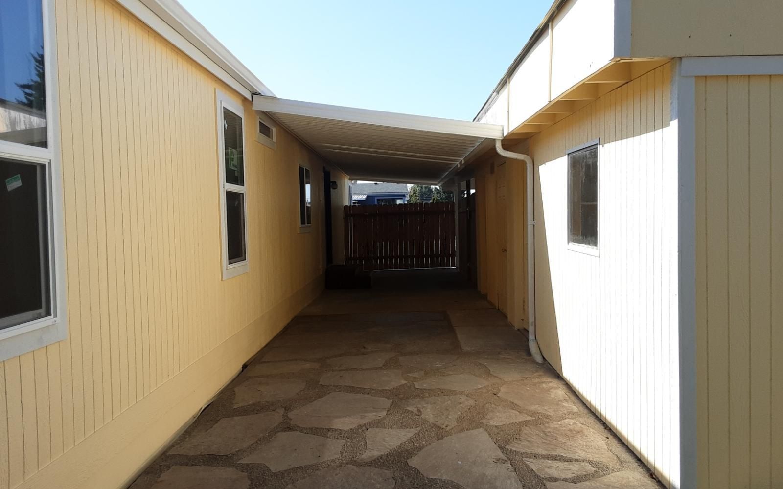 Covered walkway between beige and white buildings with a stone-patterned floor, leading to a shaded gate.