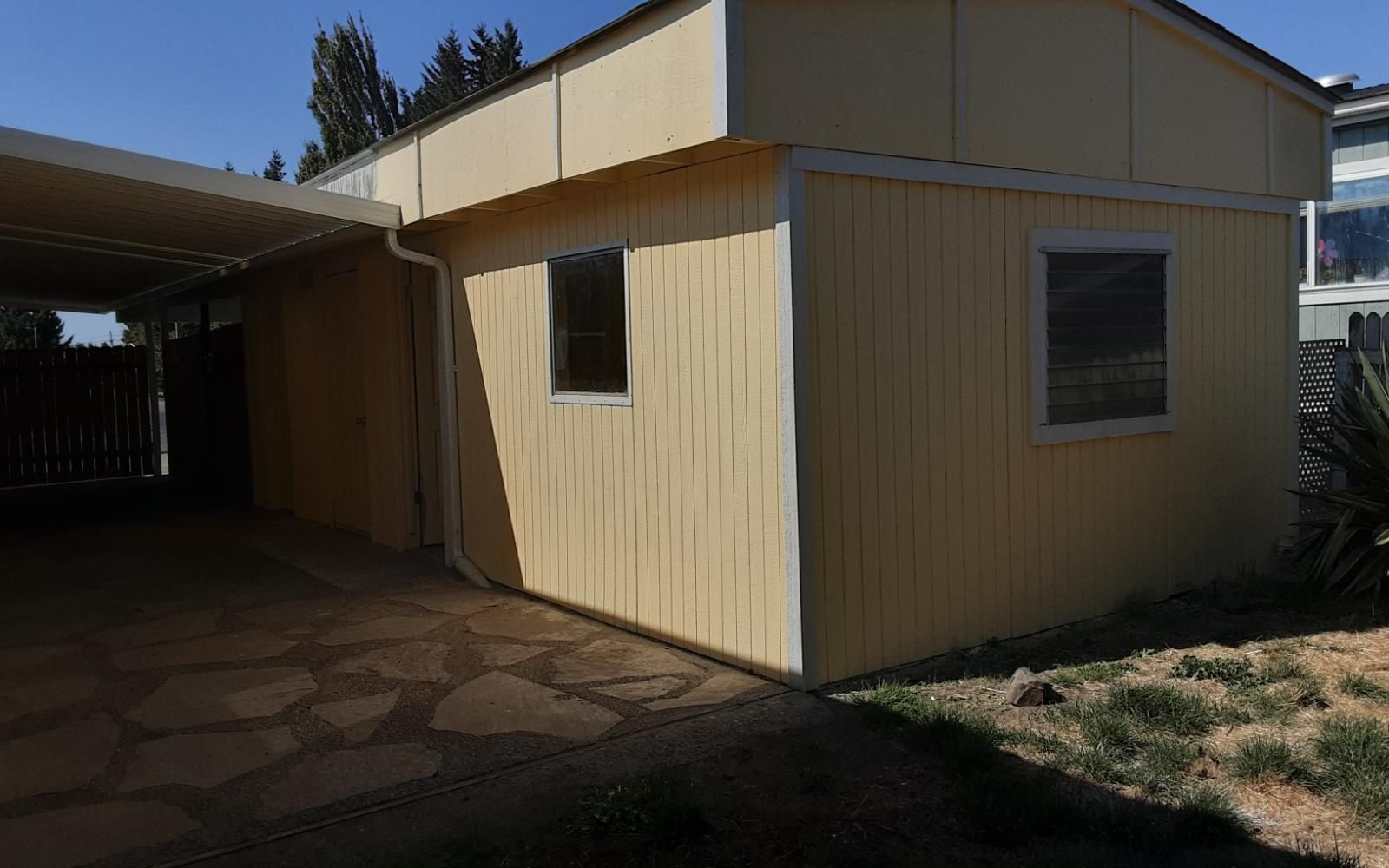 Small tan house exterior with a carport, windows, and a paved walkway in sunlight