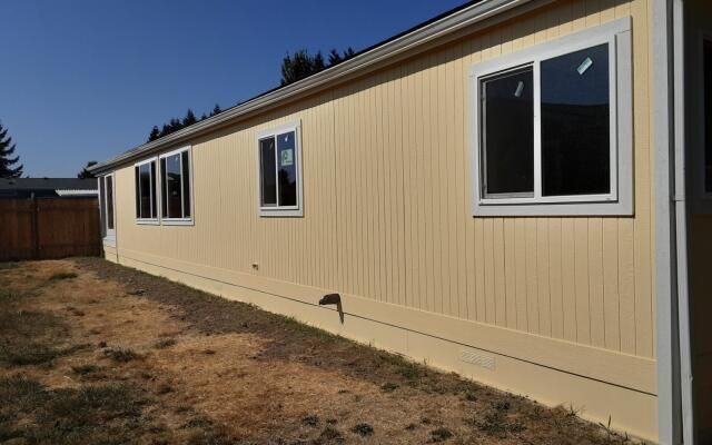 Beige manufactured home exterior with white-trimmed windows and a small dirt yard under a blue sky