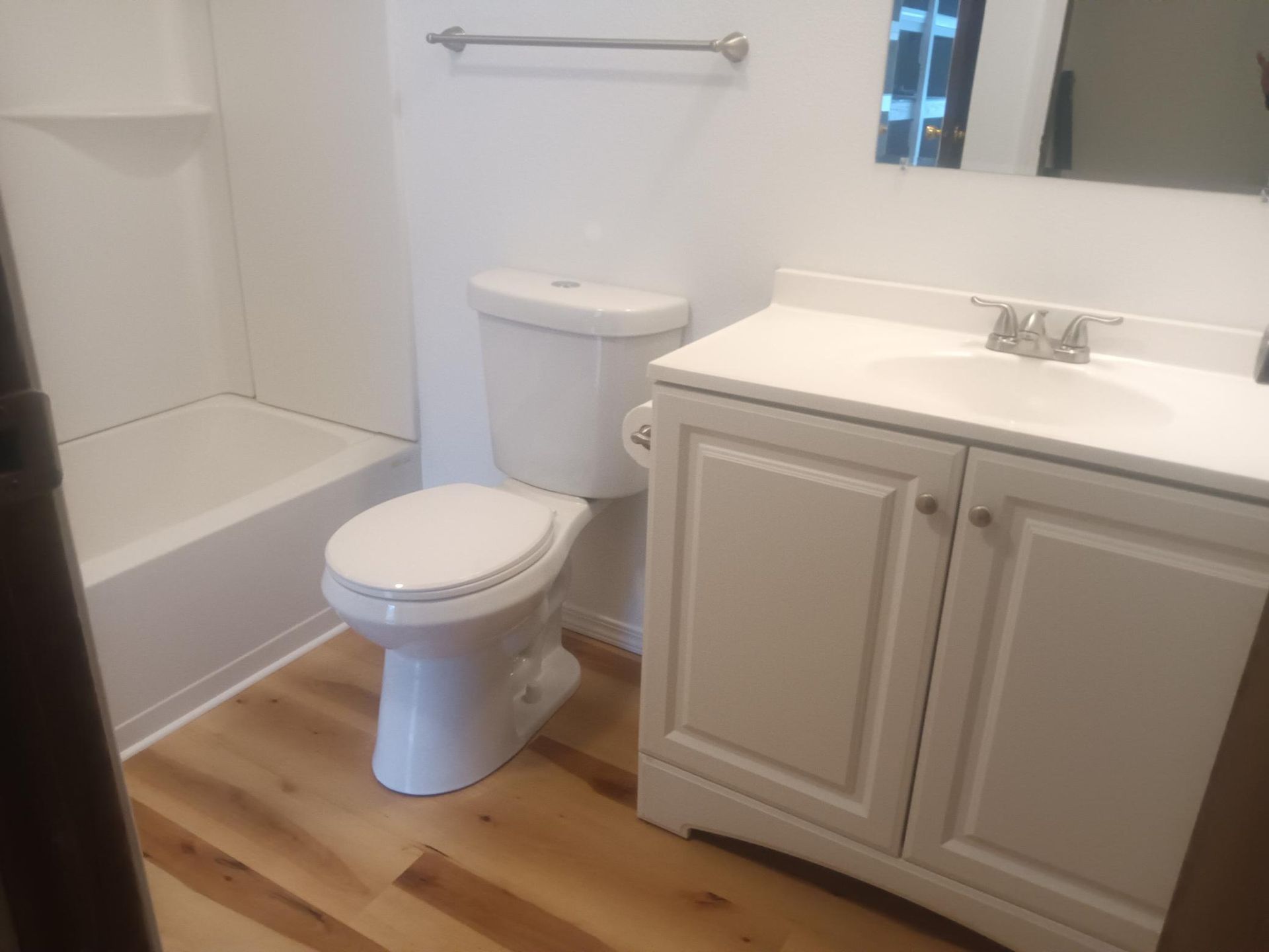 Small bathroom with toilet, tub, and white vanity on wood flooring.